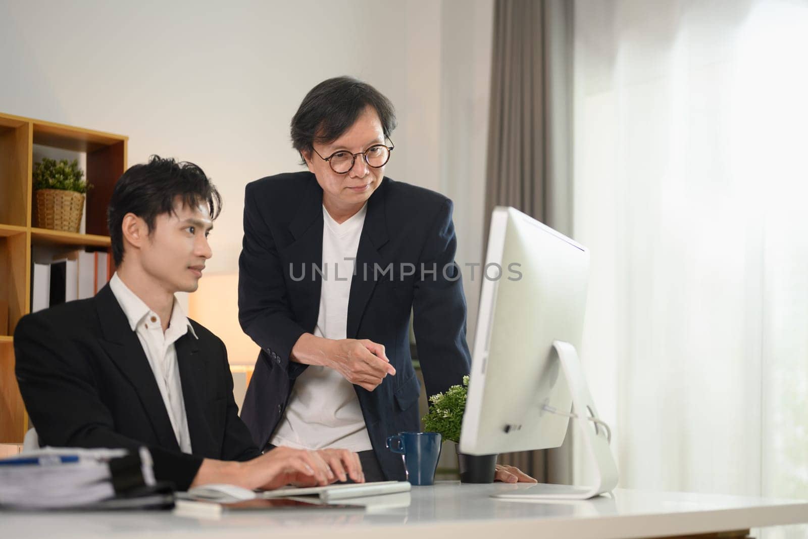 Confident middle age businessman explaining data on computer monitor to younger colleague by prathanchorruangsak