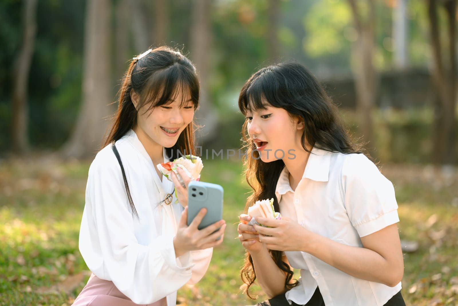Happy female friends eating sandwich and checking news feed or messaging mobile phone at outdoor.