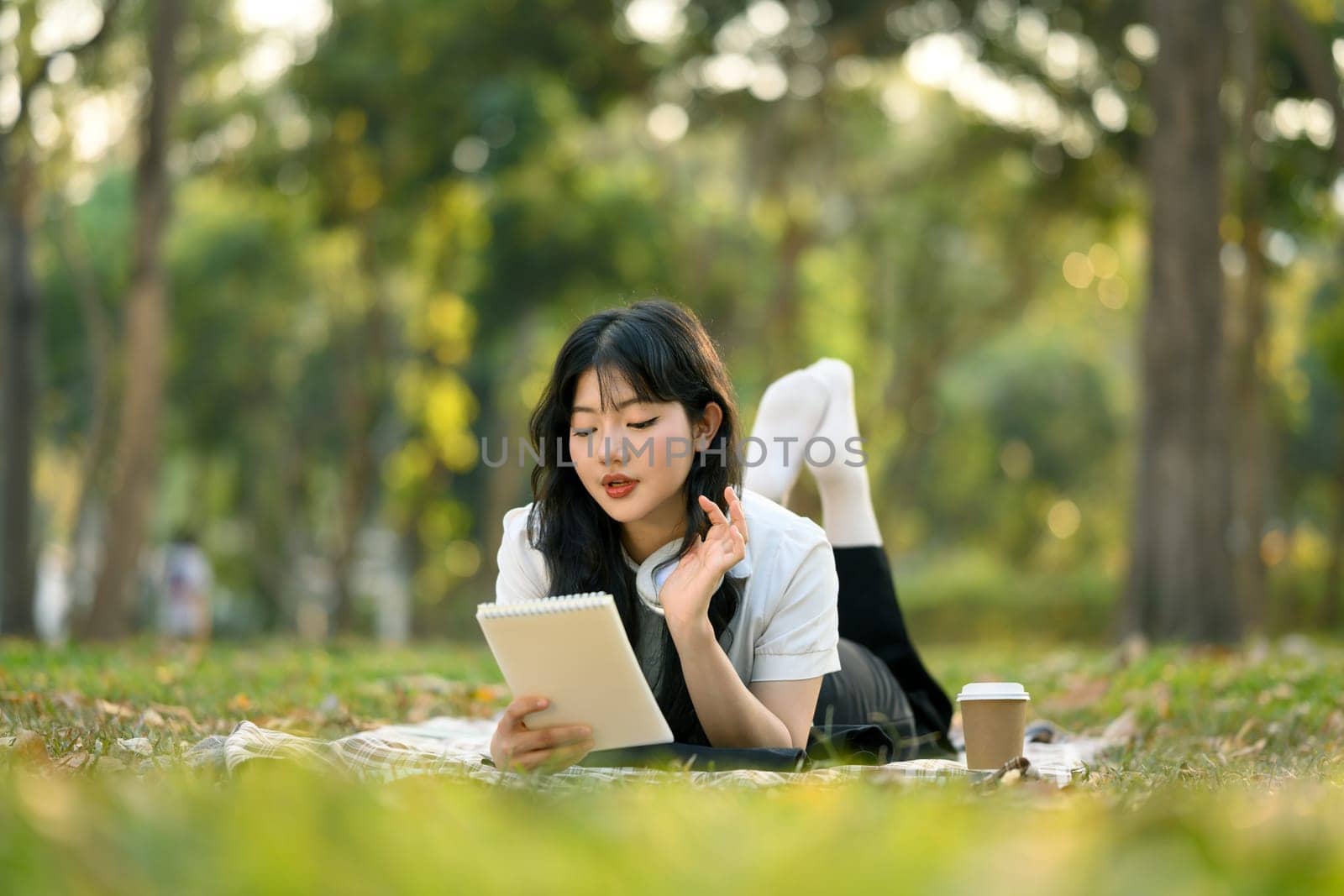 Calm Asian girl in casual clothes lying on picnic blanket and reading notes in notebook.
