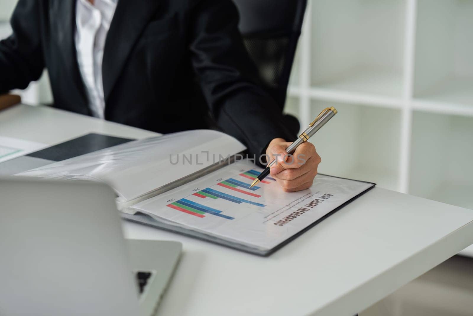 A woman is writing on a piece of paper with a pen. She is wearing a black suit and is sitting at a desk. The paper has a lot of numbers and calculations on it