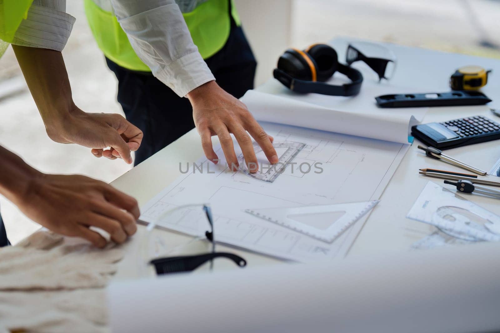 Two people are working on a blueprint, one of them is wearing a safety vest. Concept of collaboration and focus as the two individuals work together to create a detailed plan