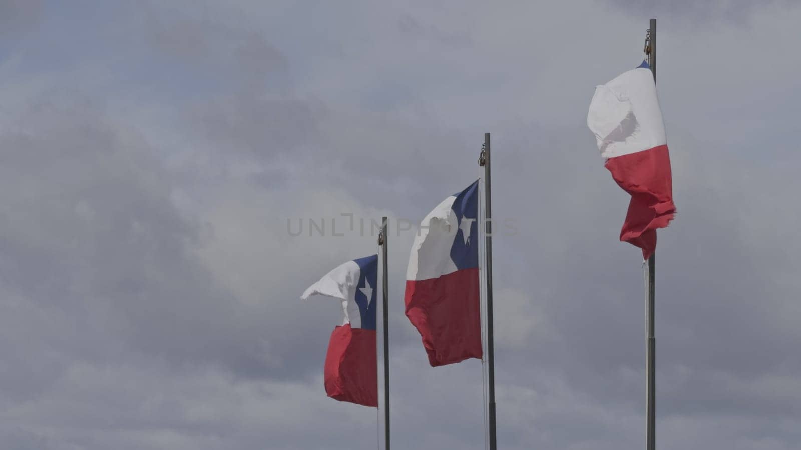 Three Chilean flags wave in the wind in slow-motion against a clear sky, positioned at different heights.