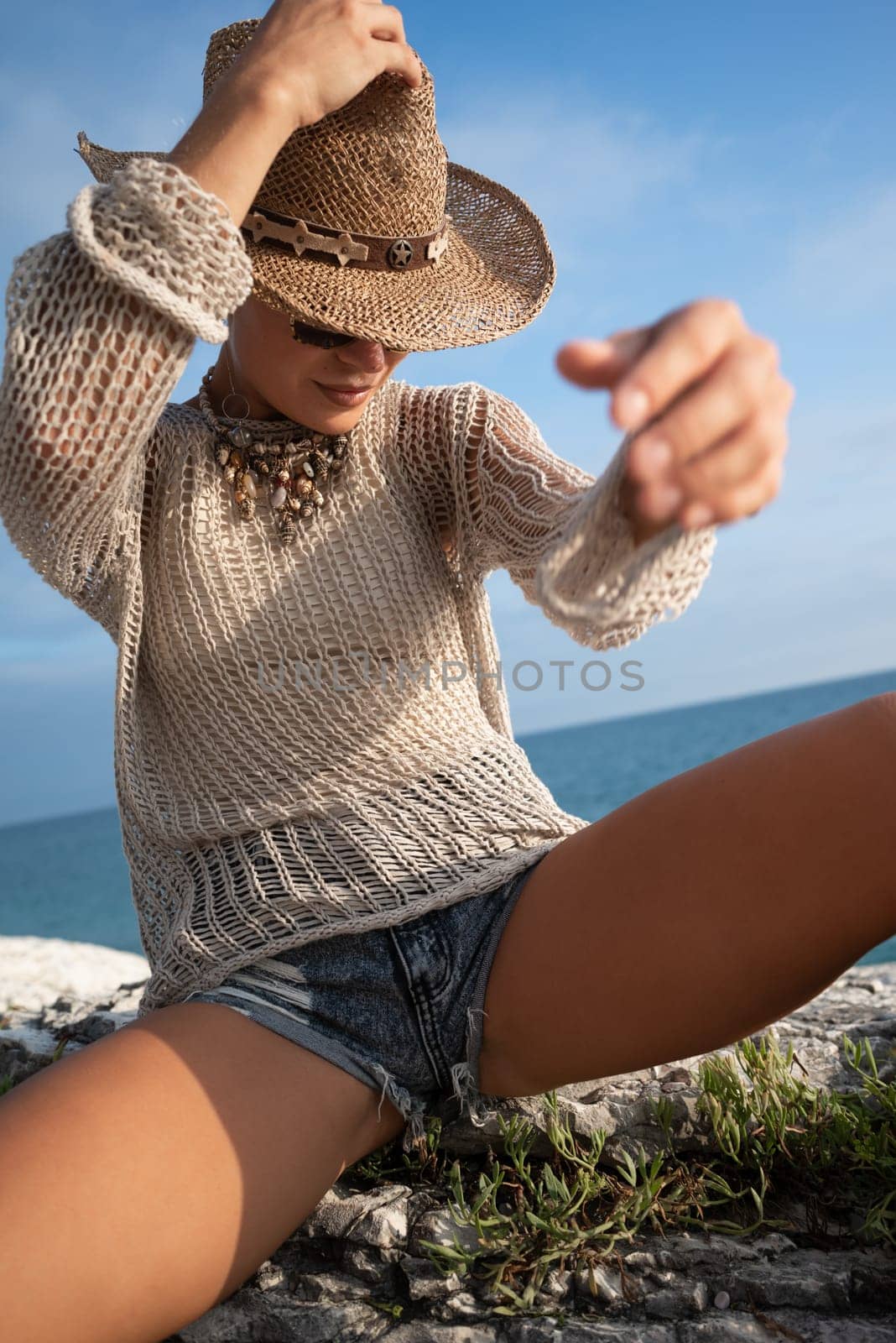 sexy girl in stylish fashionable summer clothes, cowboy hat and cool boots, posing freely on white rocks against the background of the sea by Rotozey