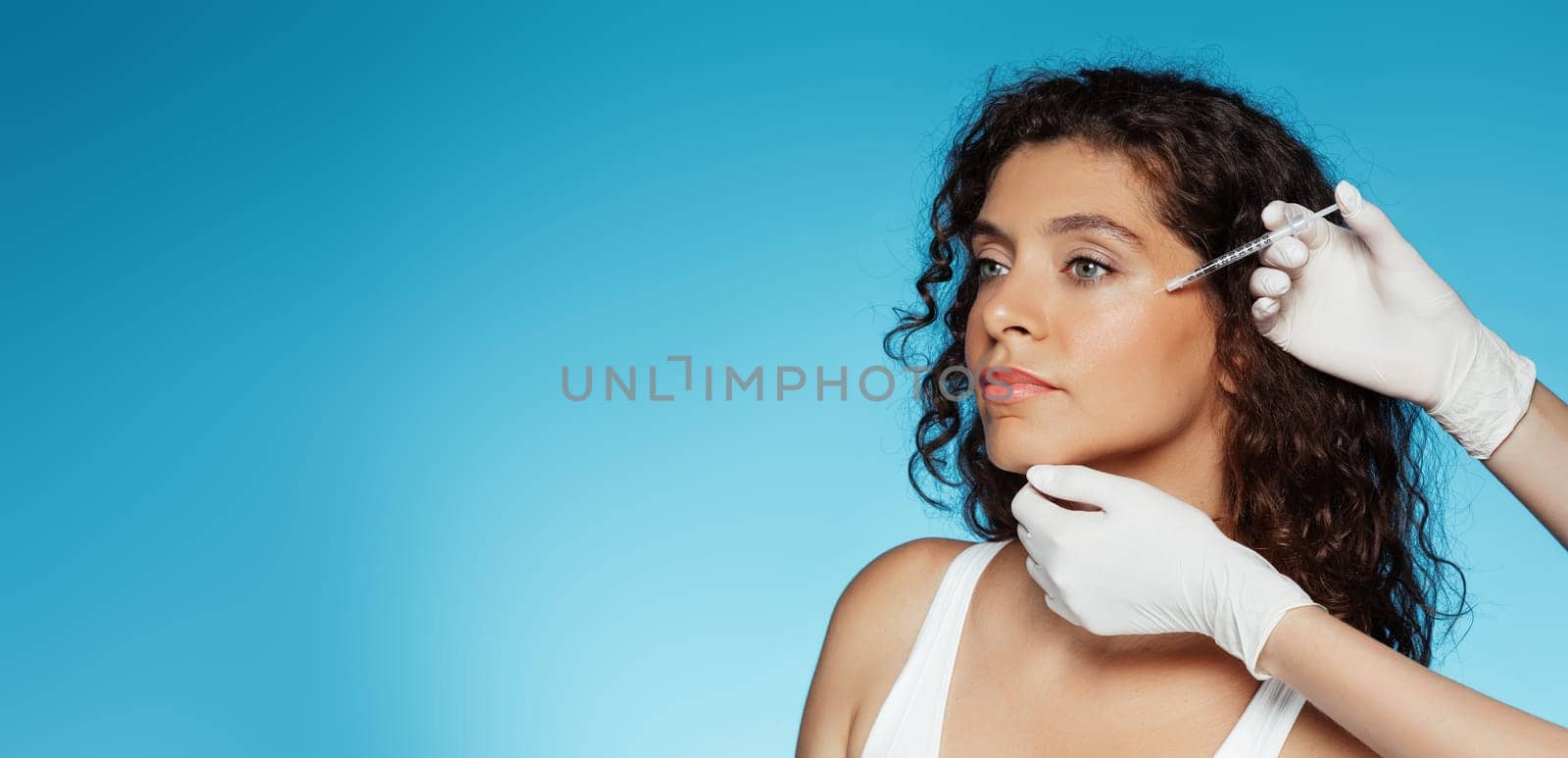 Hands of beautician making injection for calm millennial caucasian woman, isolated on blue studio background, panorama. Medical beauty care, cosmetology, anti-wrinkle procedures