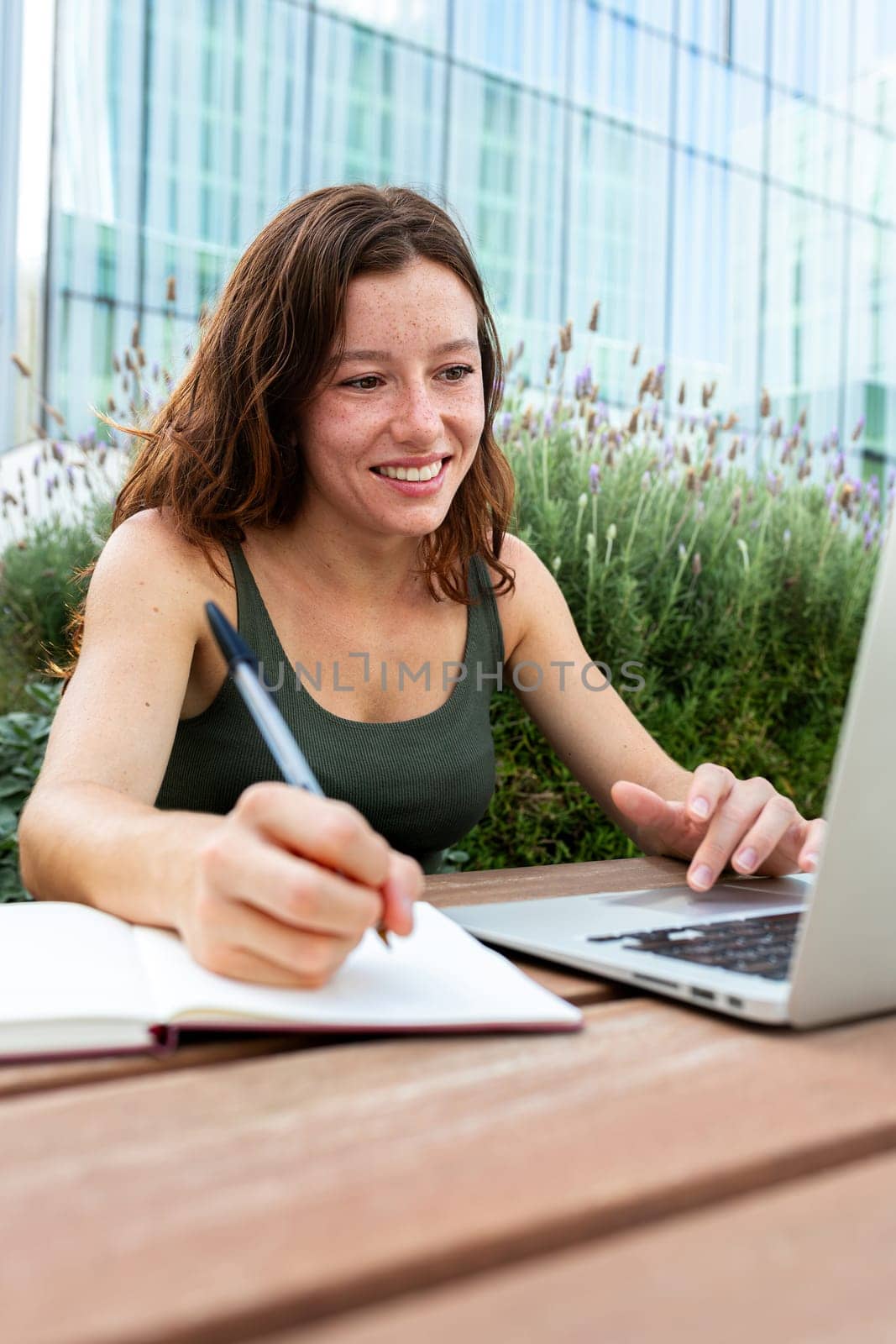 Smiling young redhead female university student writing on notebook with pen while doing homework using laptop outdoors. Vertical image. College concept.