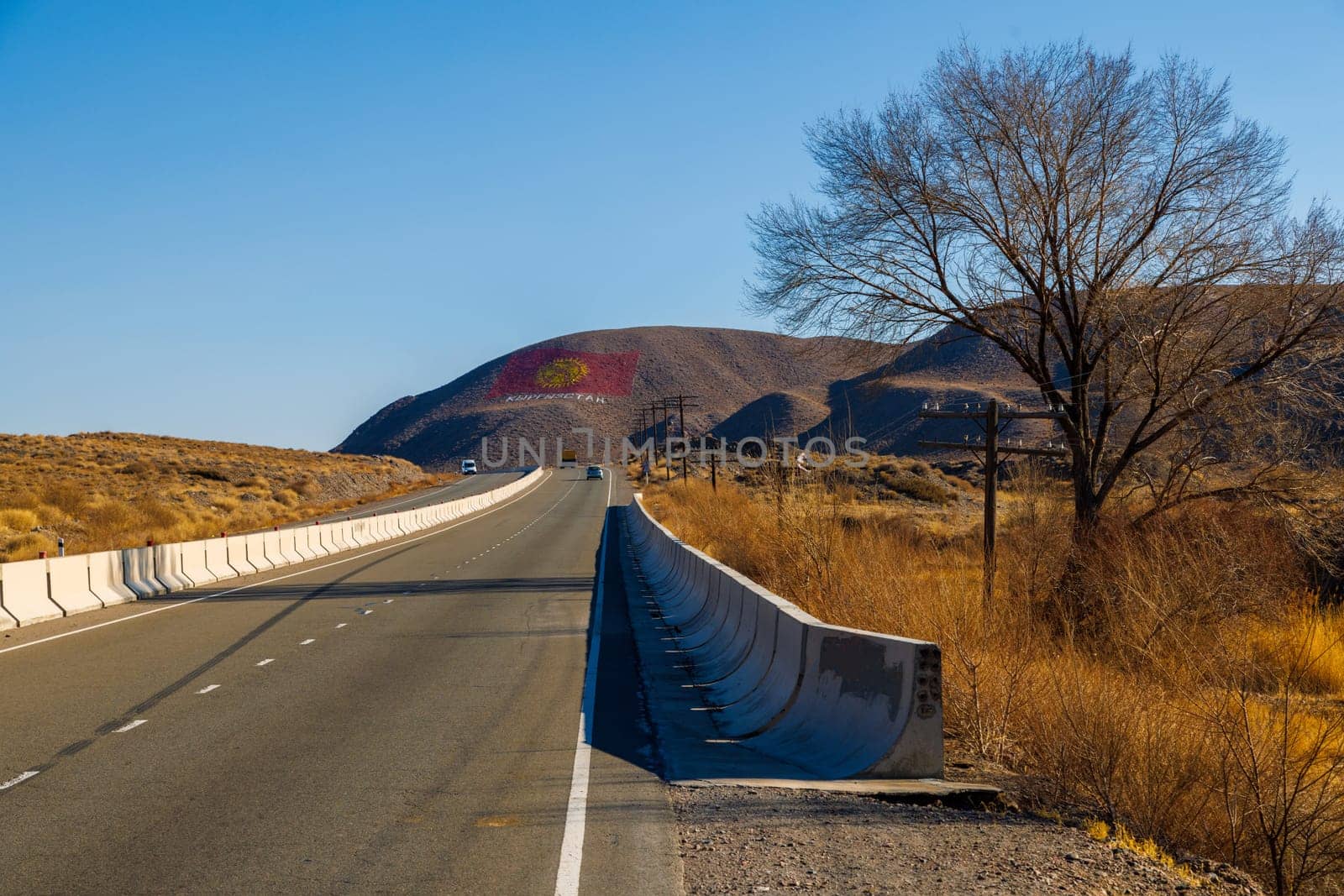 A road runs alongside a concrete barrier, dividing the asphalt from the natural landscape. The flag of Kyrgyzstan is drawn on a mountain at sunny autumn day.