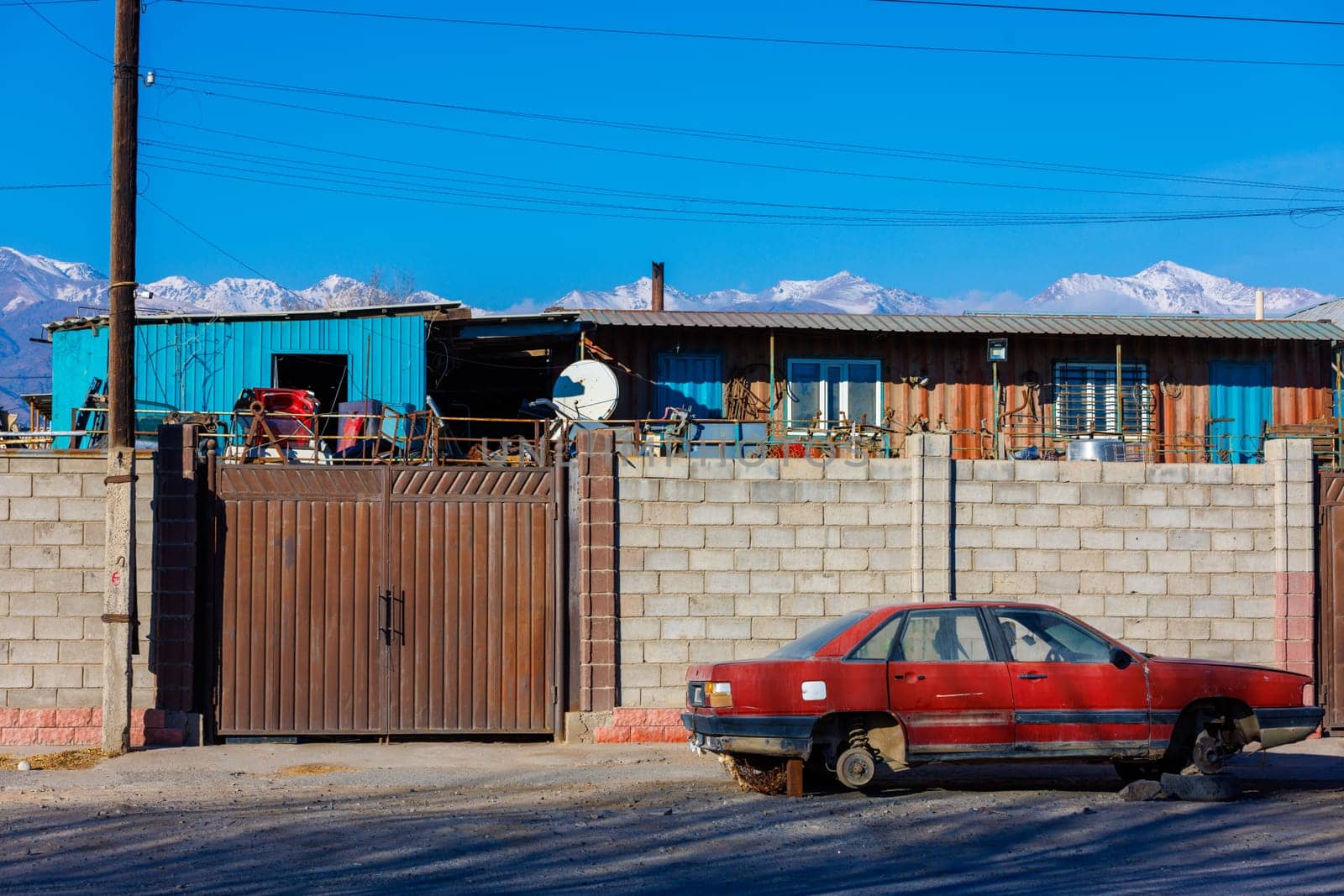 Red junked car without wheels in front of brick fence of junkyard, outside at sunny afternoon.