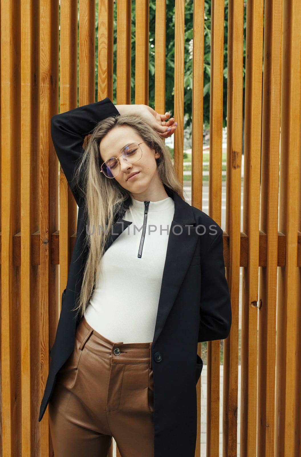 A fashionably dressed woman with glasses poses with one hand on her head, standing in front of a vertical wooden slat fence.