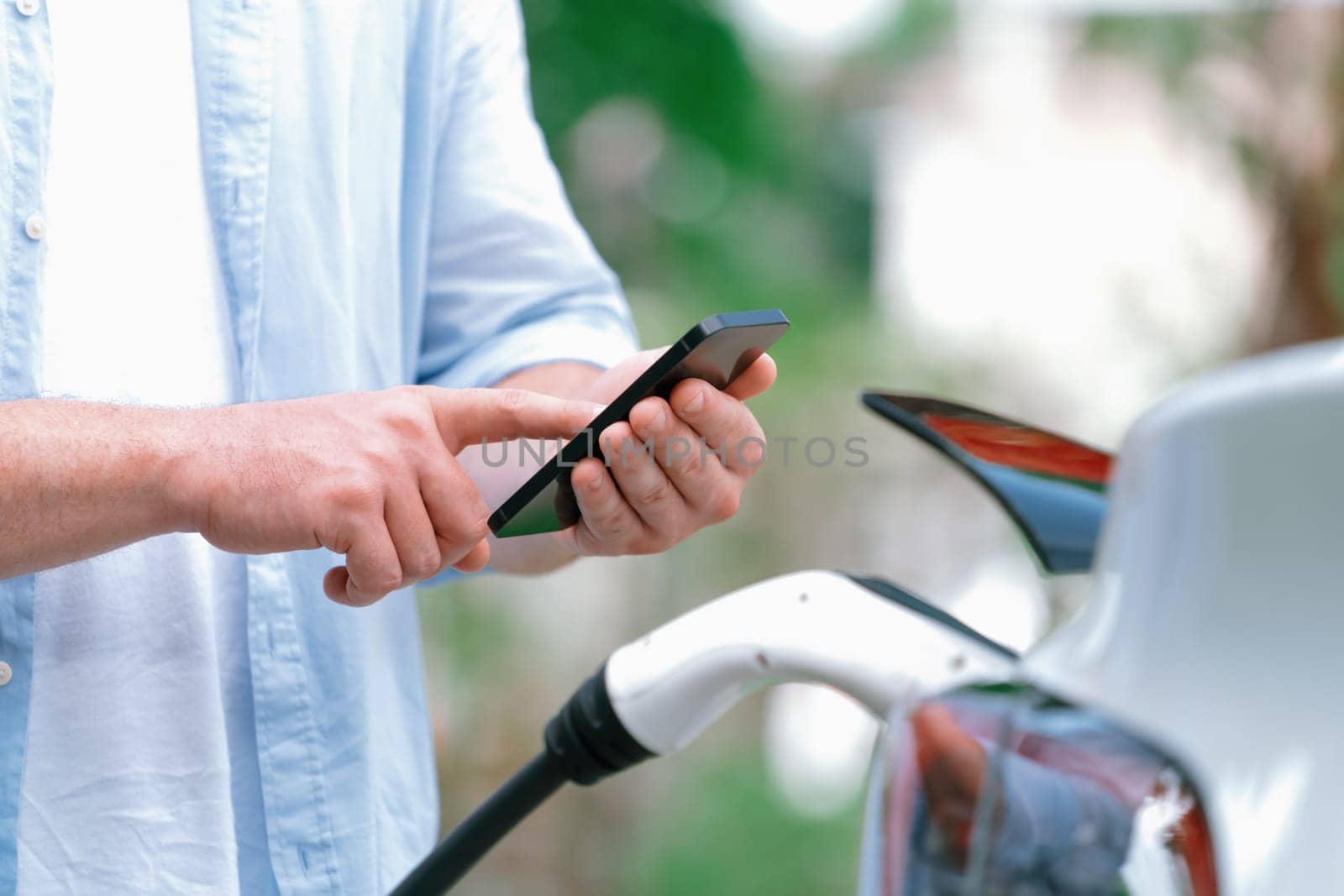 Modern eco-friendly man recharging electric vehicle from EV charging station, using Innovative EV technology utilization for tracking energy usage to optimize battery charging on smartphone.Synchronos