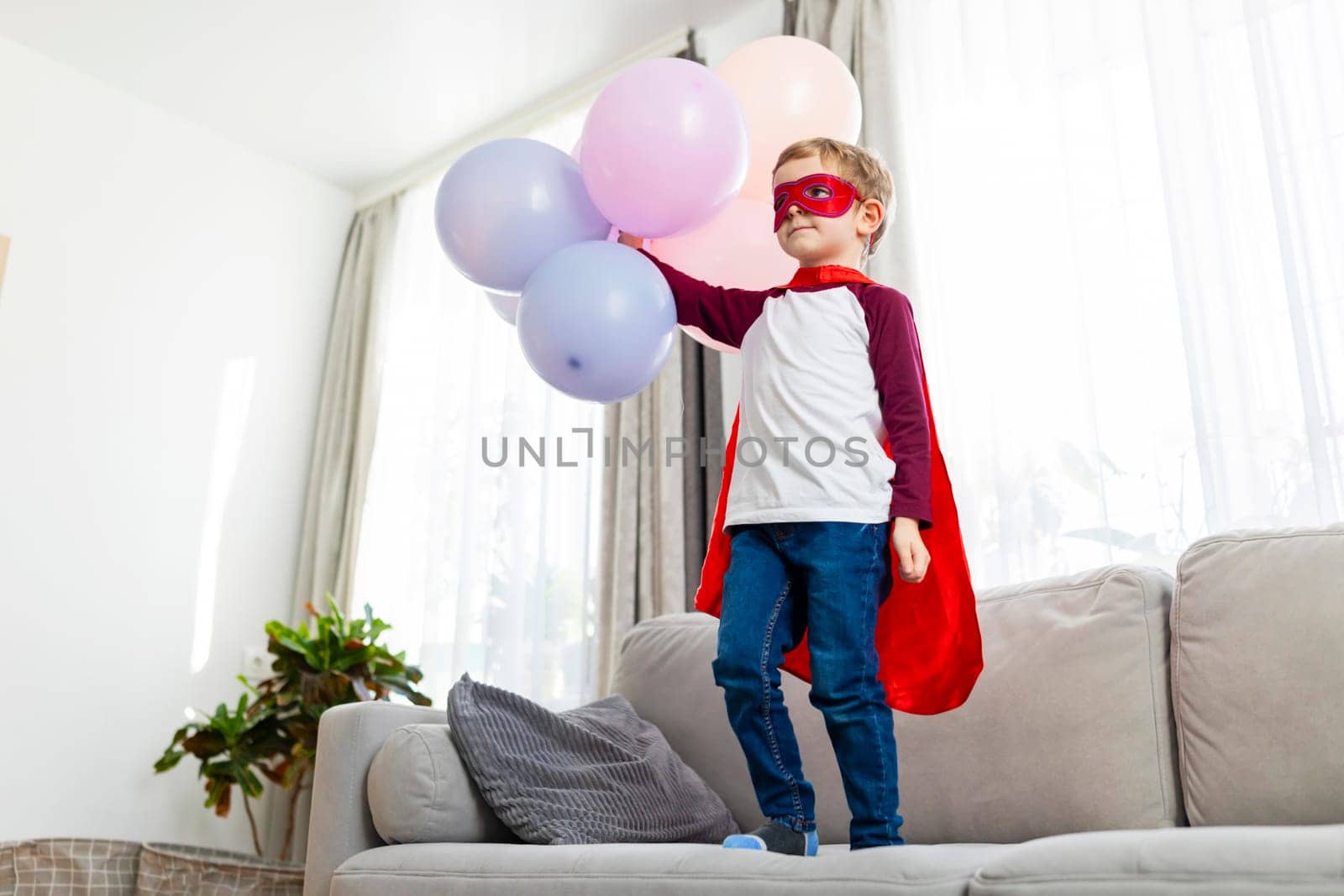 Boy in superhero costume with balloons standing on sofa. Indoor childhood play and imagination concept for design and print
