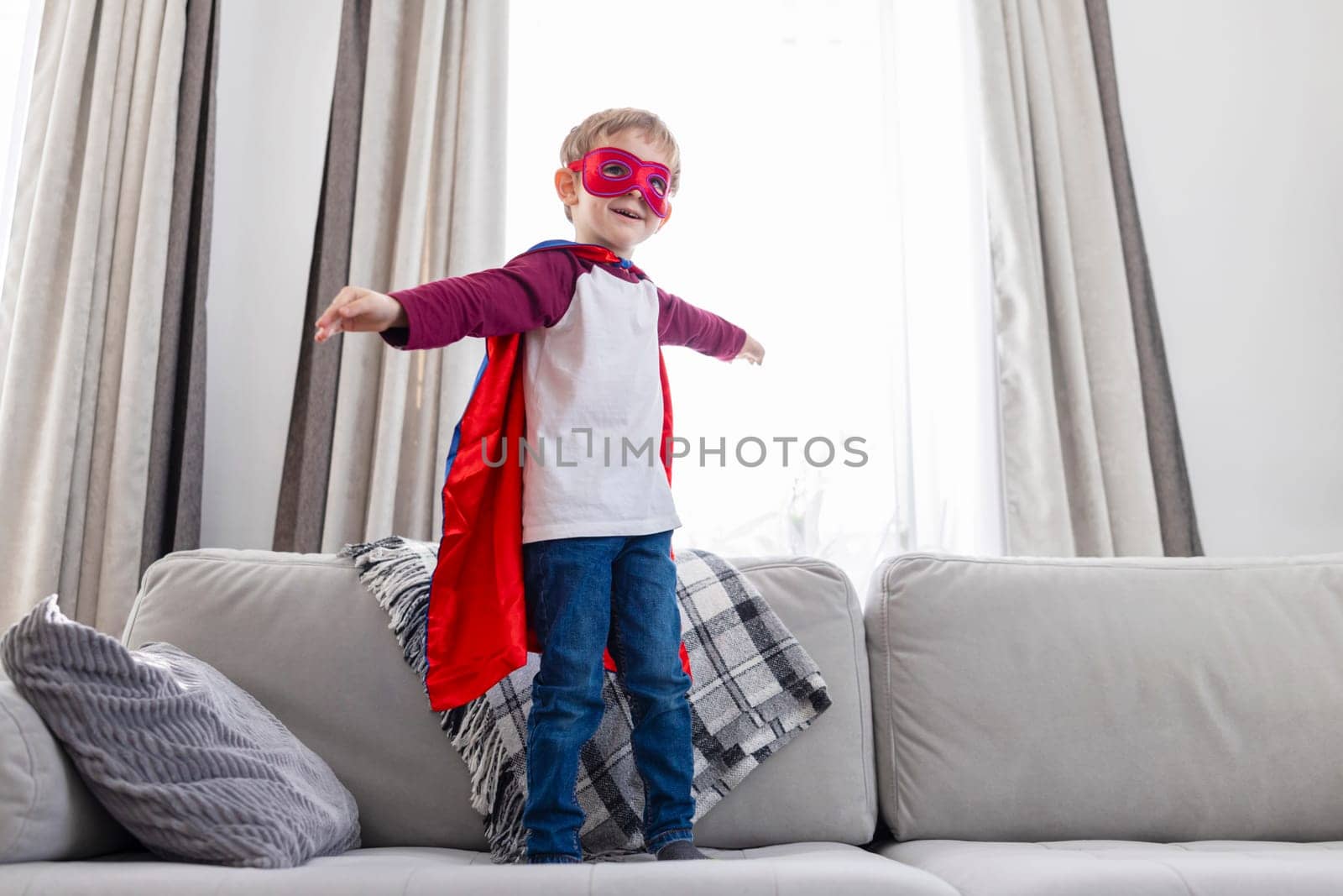 Boy in superhero costume with red cape and mask standing on couch. Indoor leisure and play concept. Design for poster, children's book illustration