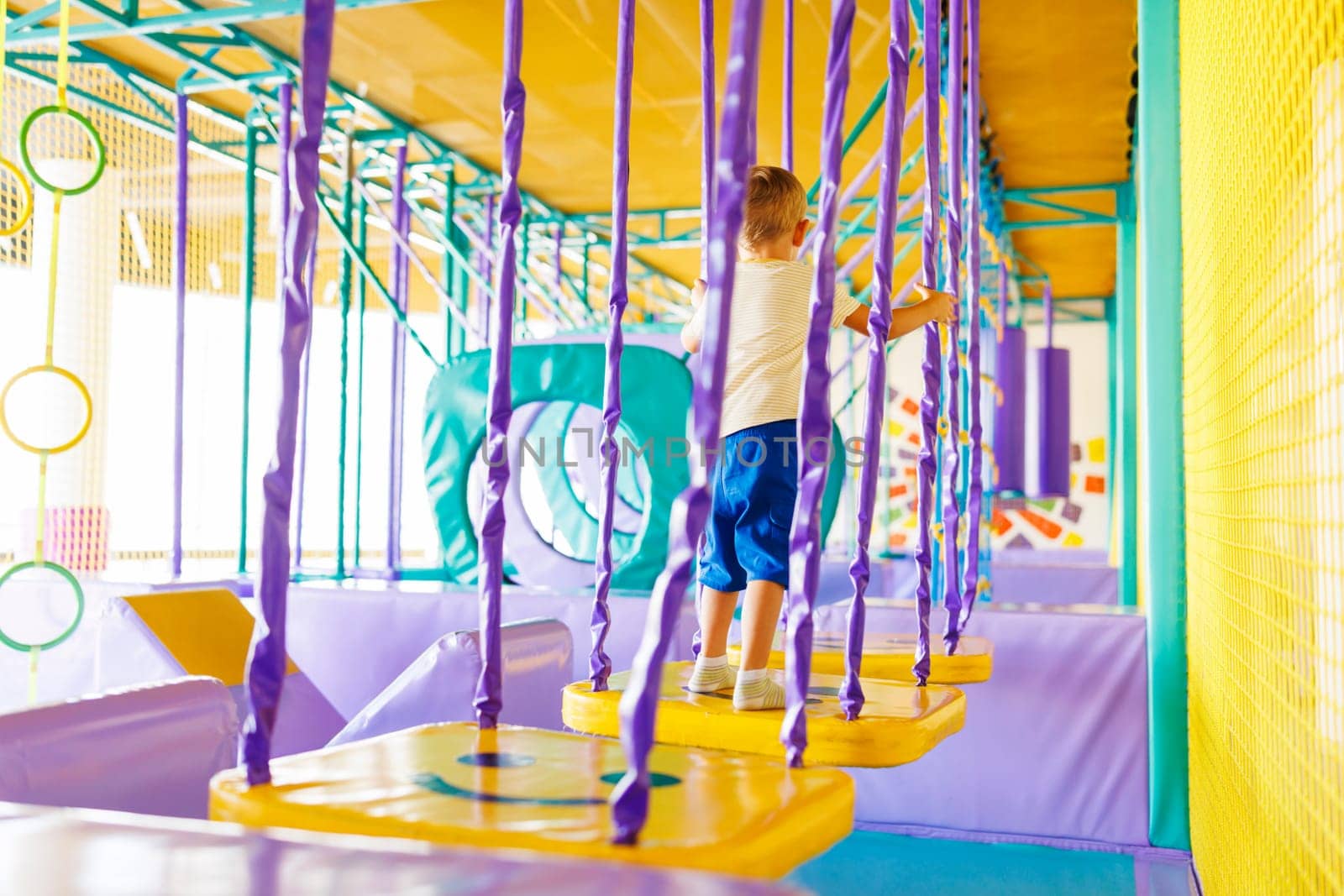 Child navigating through purple and yellow play structure in indoor playground. Brightly colored play area for kids with obstacle course. Child activity and playtime concept for design and print. Indoor entertainment