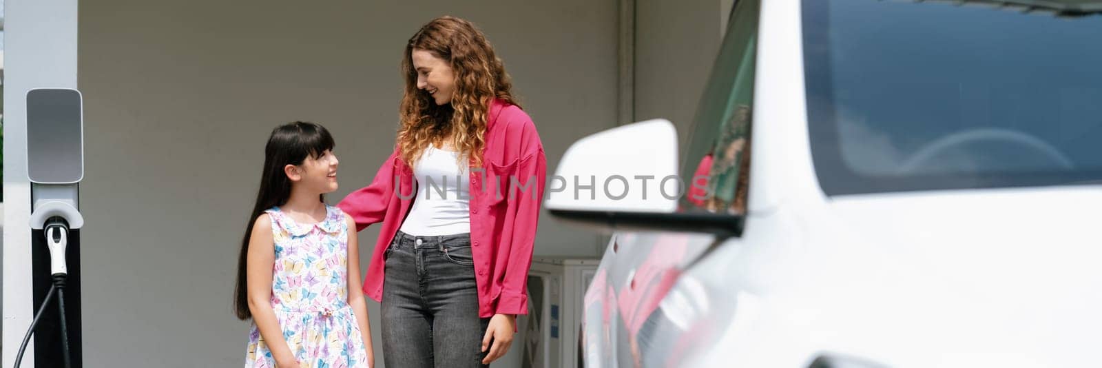 Happy little young girl learn about eco-friendly and energy sustainability as she help her mother recharge electric vehicle from home EV charging station. EV car and modern family. Panorama Synchronos