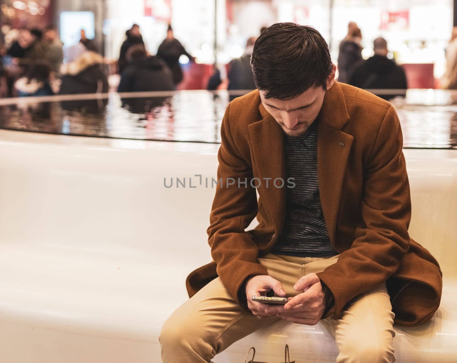 One young handsome caucasian brunette guy in a brown coat and beige trousers reads an article in a smartphone while sitting on a plastic white bench with a fountain in a store in a shopping center, close-up top view. The concept of using technology, online shopping.
