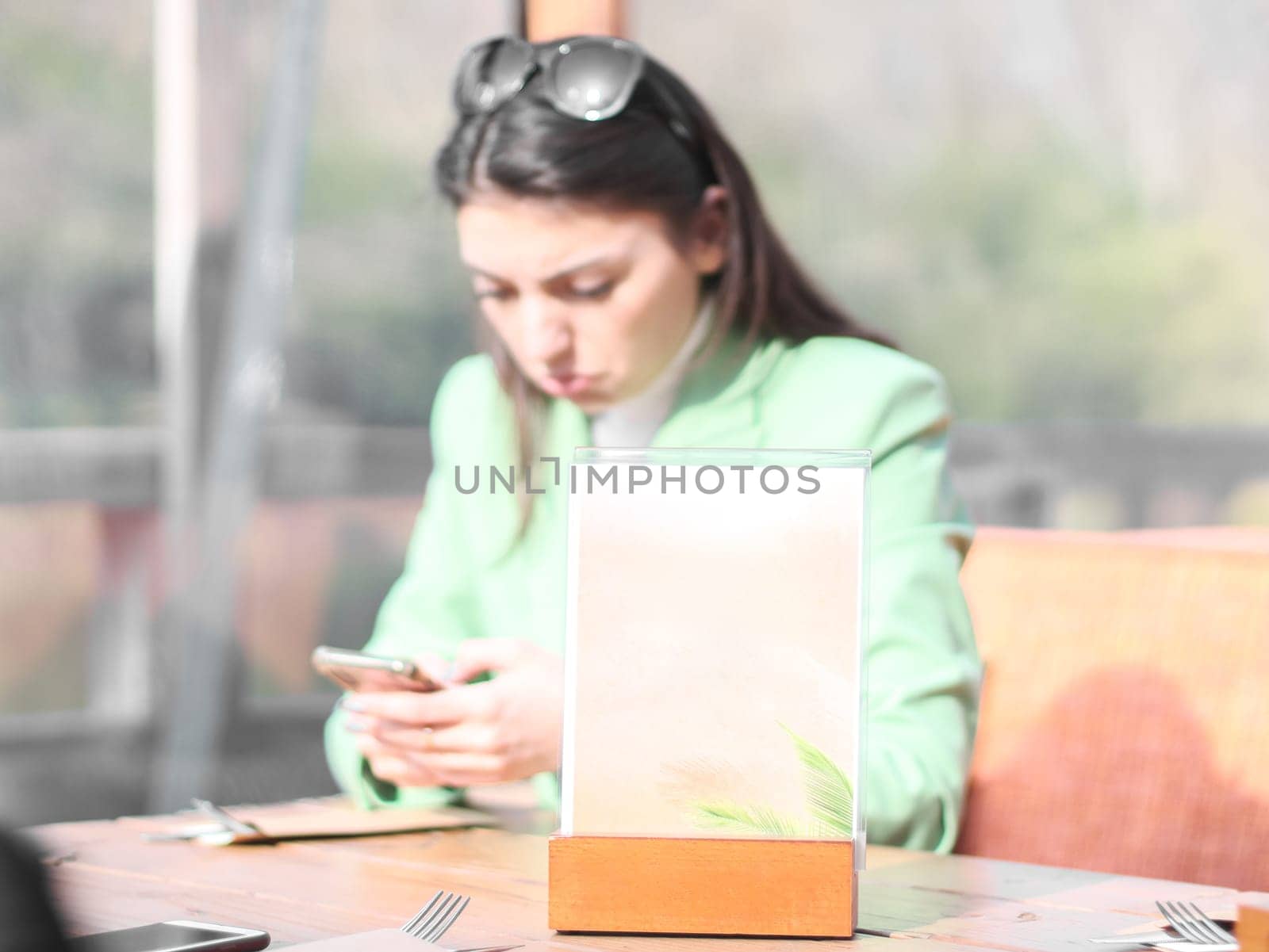 One empty sign for a restaurant menu stands on the table with a blurred sad and disappointed girl with a mobile phone in her hands who is sitting on the terrace of a water cafe on a sunny spring day, side view close-up with depth of field.