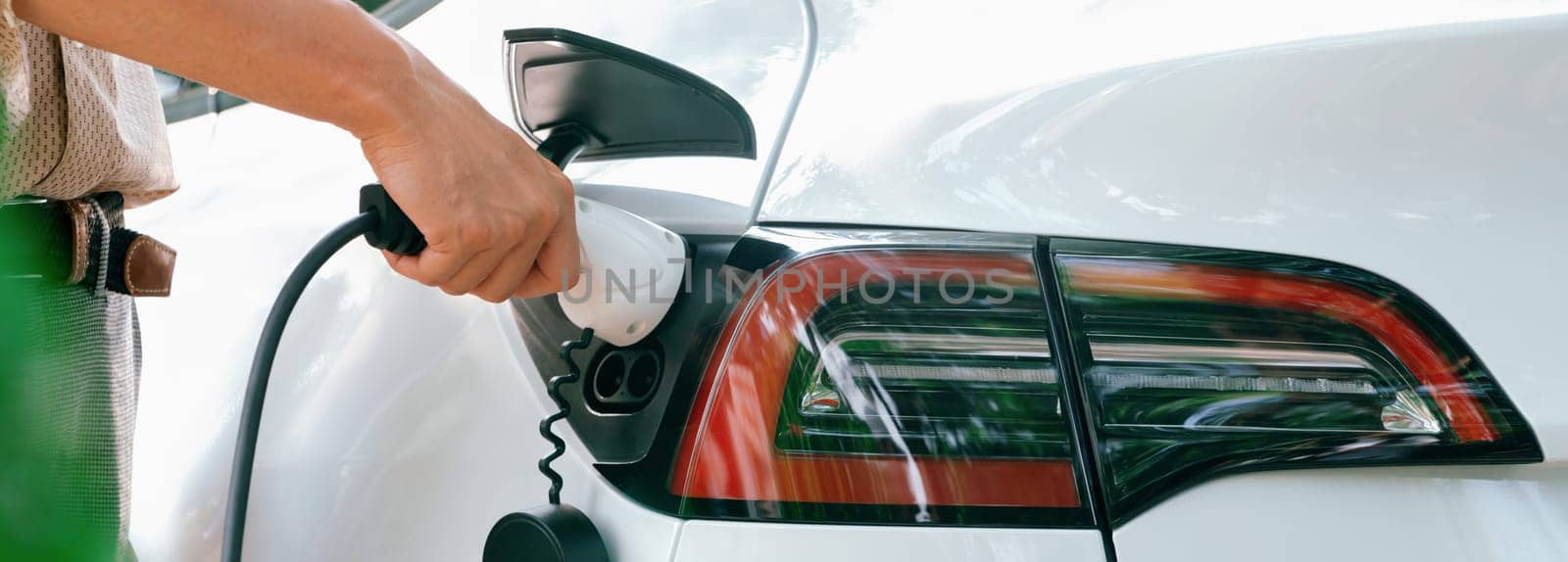 Young man recharge electric car's battery from charging station in outdoor green city park. Rechargeable EV car for sustainable environmental friendly urban travel. Panorama Expedient