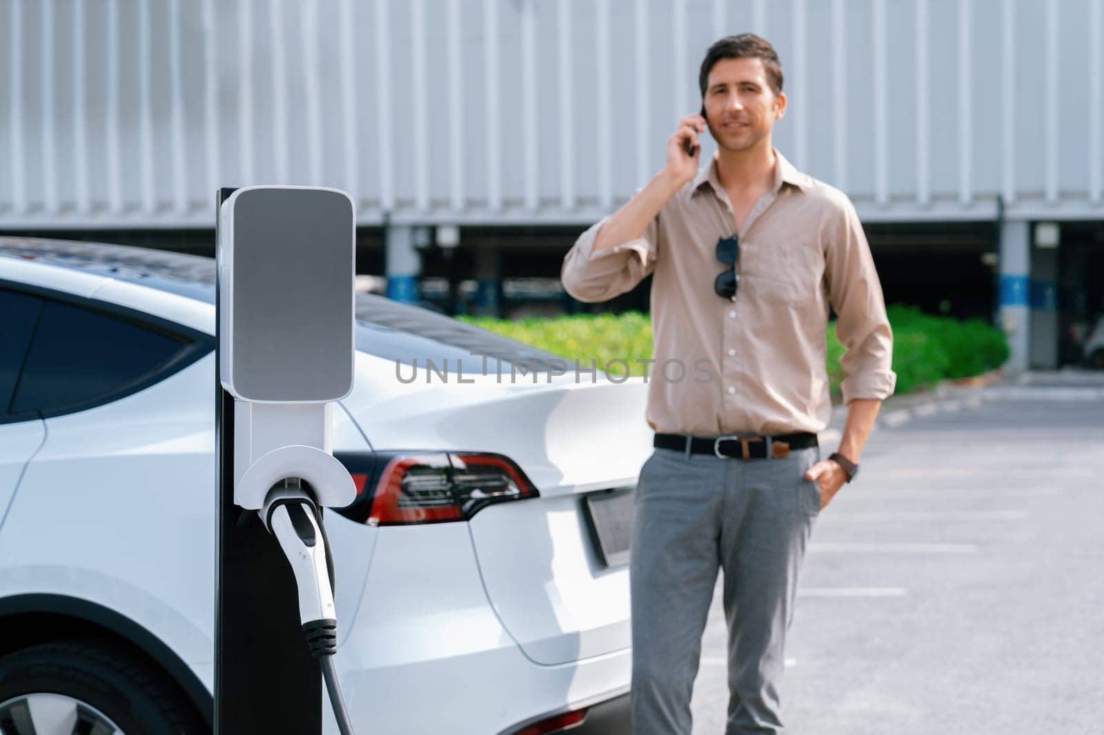 Young man recharge EV electric vehicle at green city commercial mall parking lot while talking on phone. Sustainable urban lifestyle for eco friendly EV car with battery charging station. Expedient