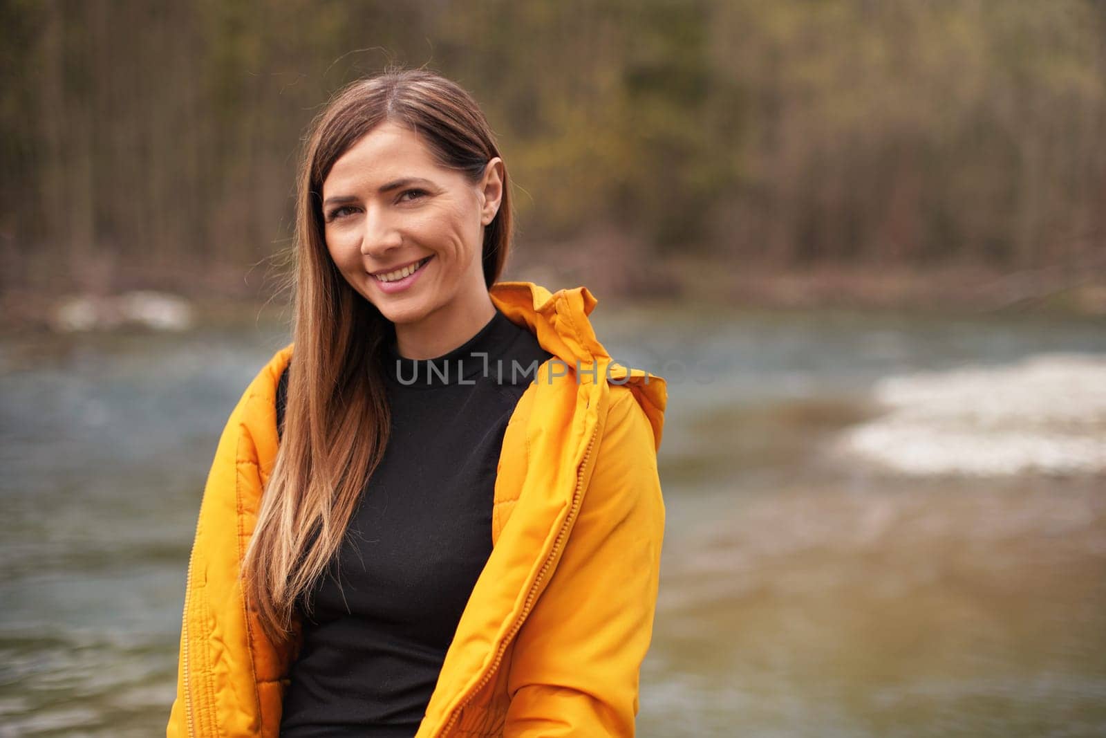 Portrait of young woman in yellow jacket, smiling, long hair down, blurred river background