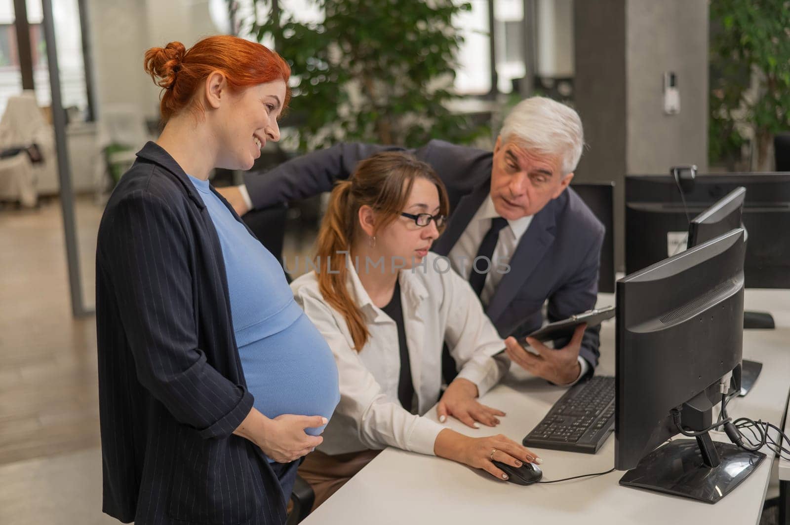 An elderly man, a Caucasian woman and a pregnant woman are discussing work issues at the computer