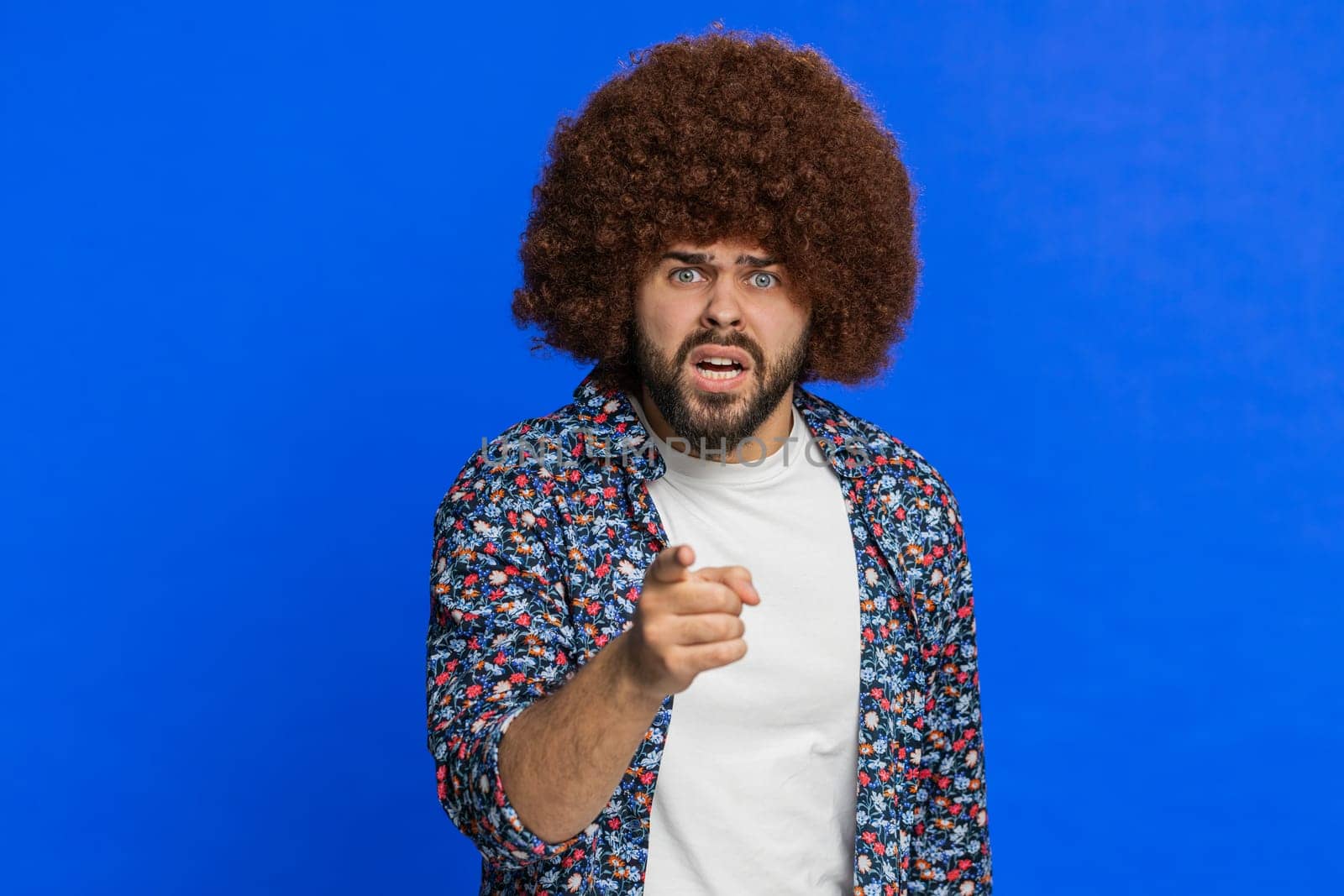 Quarrel. Displeased aggressive unhappy man with Afro hairstyle wig gesturing hands with irritation displeasure blaming scolding for failure asking why this happened. Guy isolated on blue background