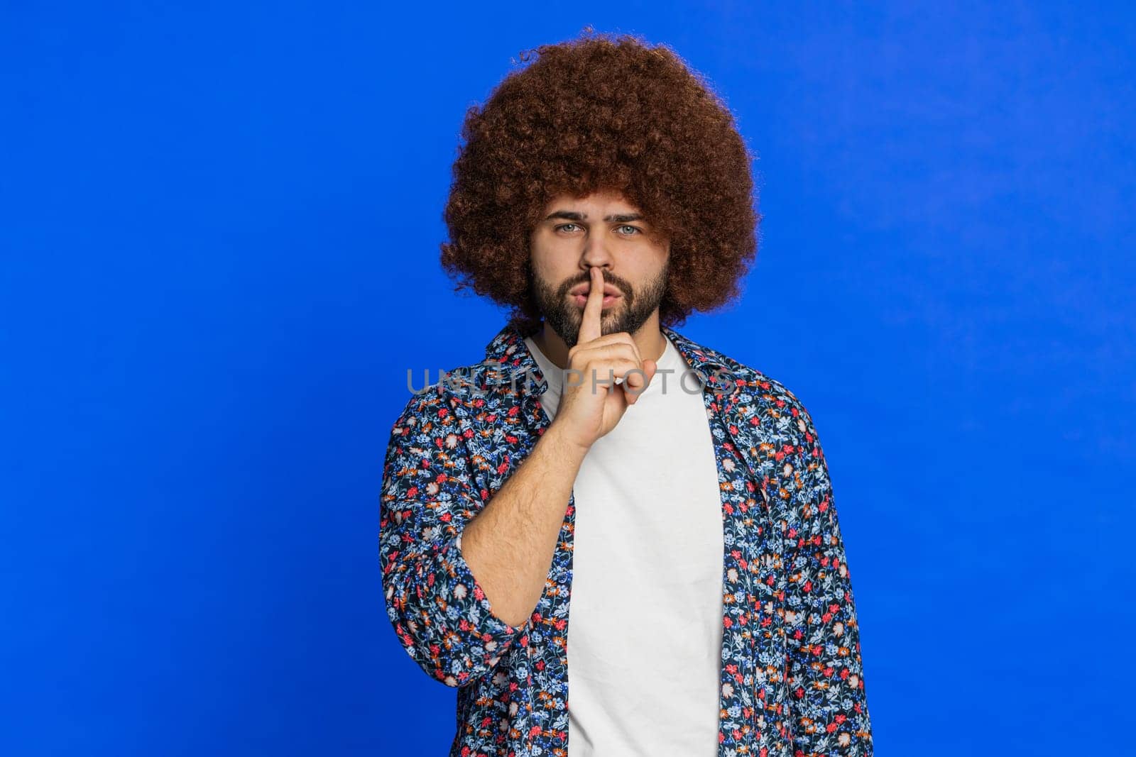 Shh be quiet please. Caucasian man presses index finger to lips makes silence gesture sign do not tells secret, stop talk gossip, confidential privacy. Stylish guy on blue background. Copy-space