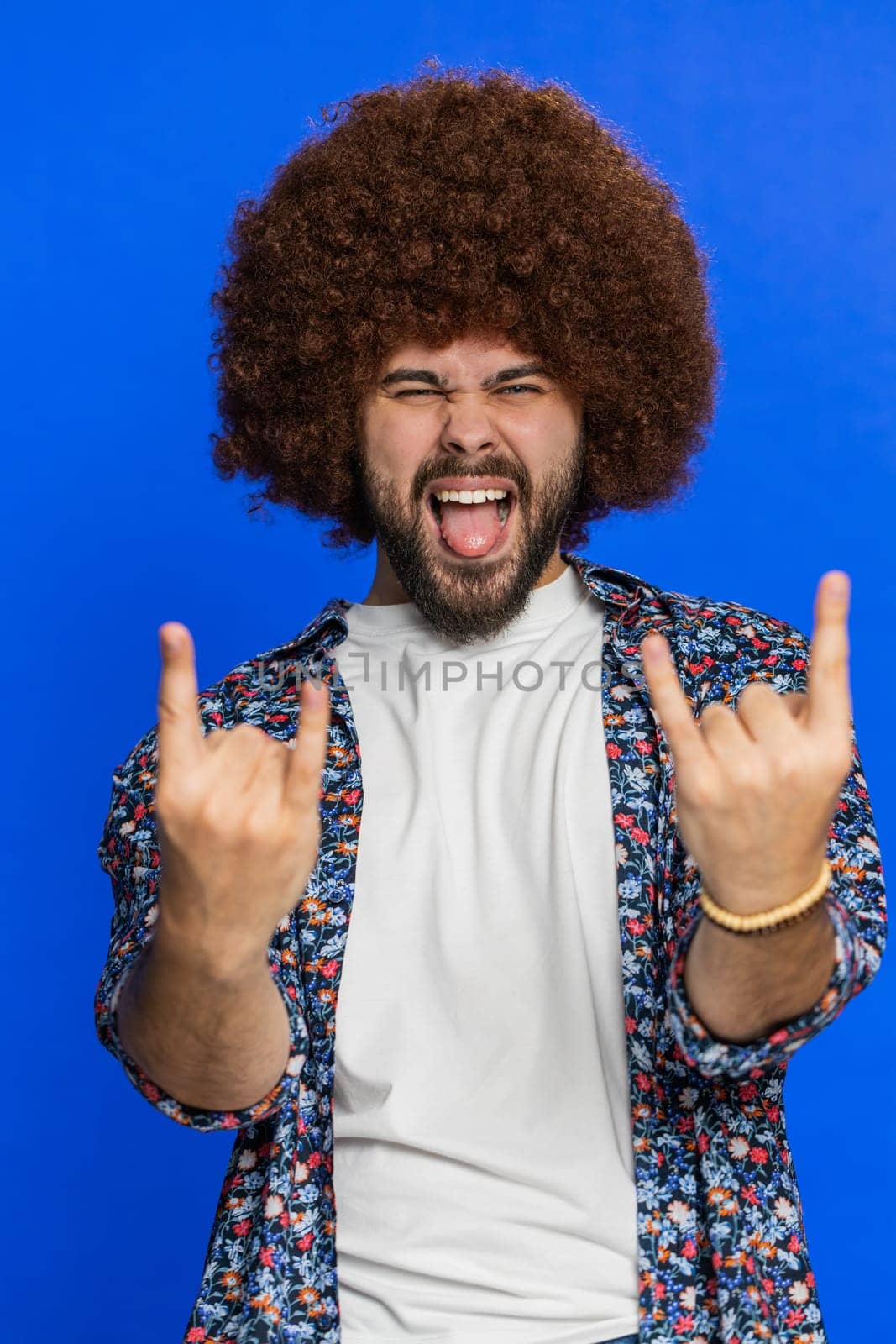 Overjoyed happy Caucasian man showing rock n roll gesture by hands, cool sign shouting yeah with crazy expression dancing rejoicing victory win success. Stylish young guy isolated on blue background