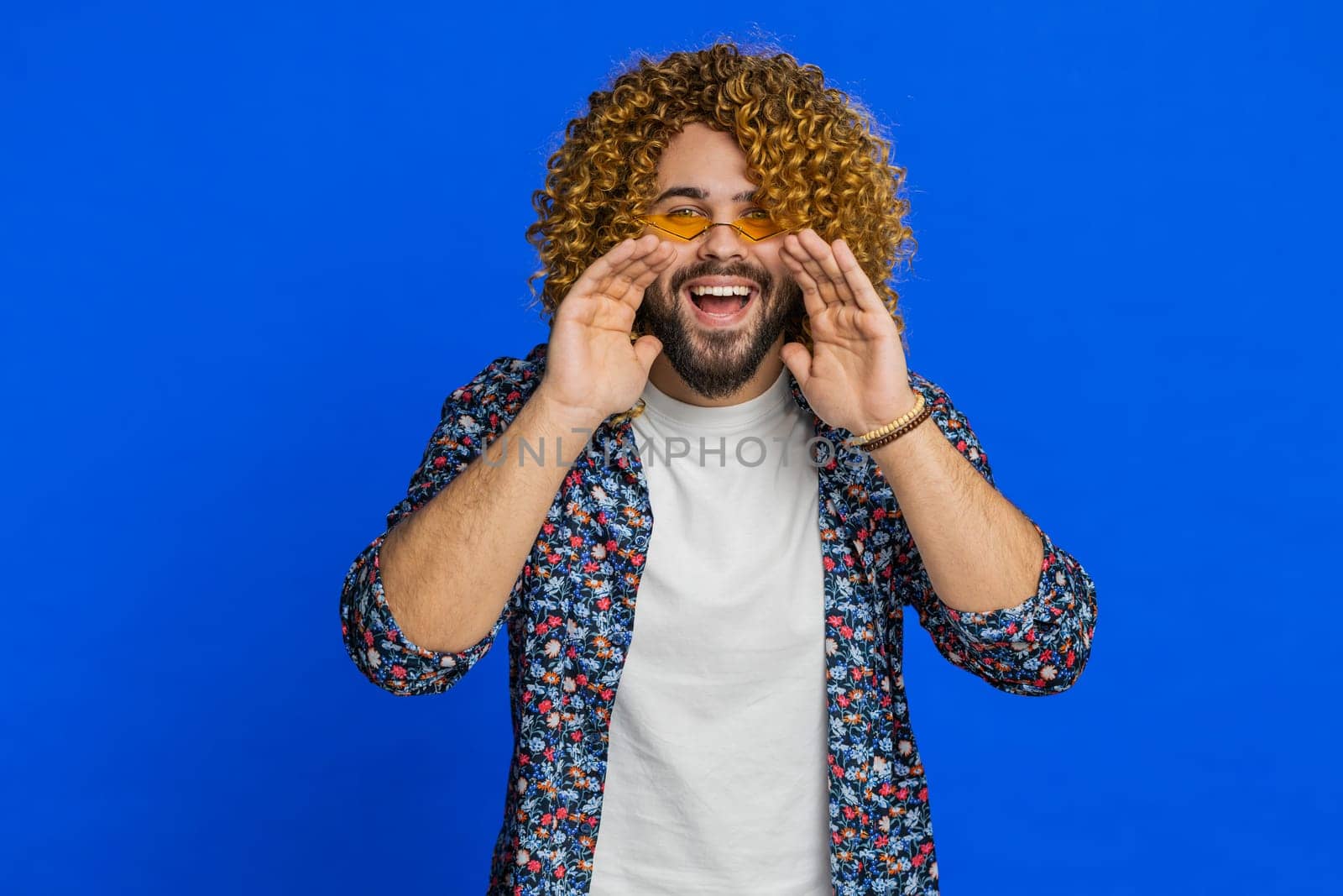 Happy excited Caucasian man yell, shout to camera, promotion advertisement of holidays sale, invitation, greetings, welcome. Excited smiling stylish young guy scream loud isolated on blue background