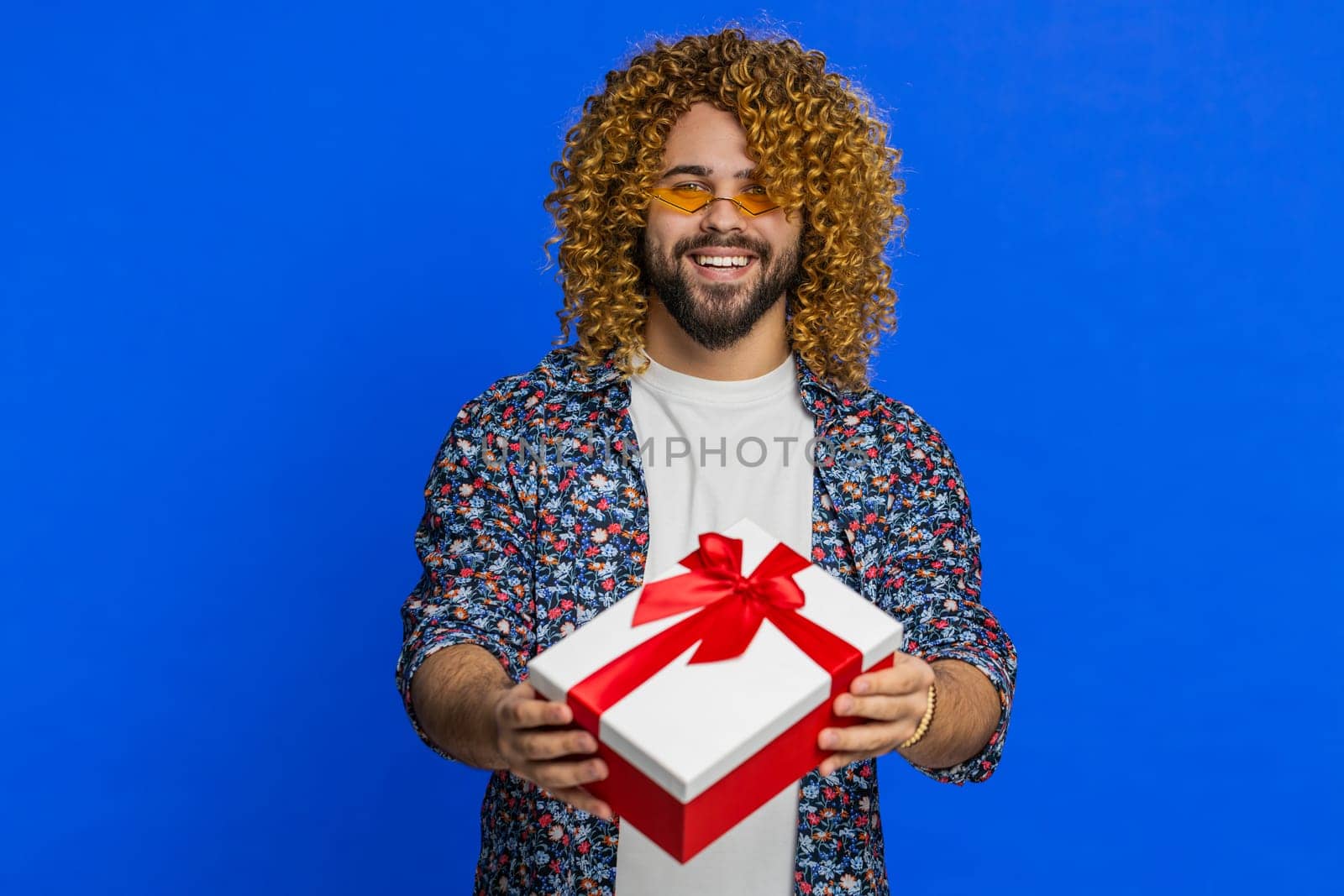 Positive smiling Caucasian man presenting birthday gift box stretches out hands offer wrapped present career bonus celebrating party promotion discount sale. Stylish guy isolated on blue background