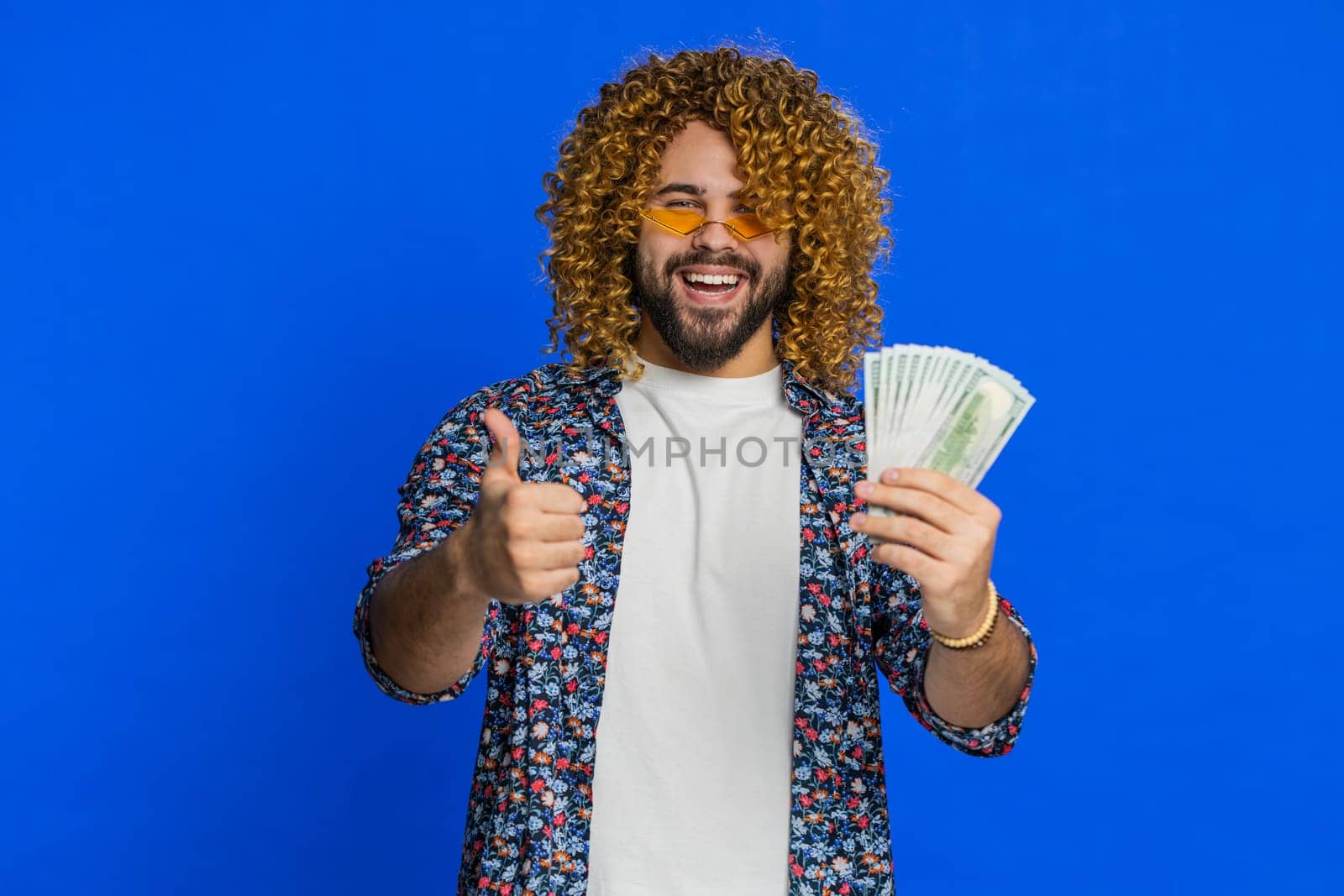 Rich excited happy man with curly hairstyle wig waving money dollar cash banknotes bills like a fan, success business career, lottery winner, big income wealth. Guy isolated on blue background indoors