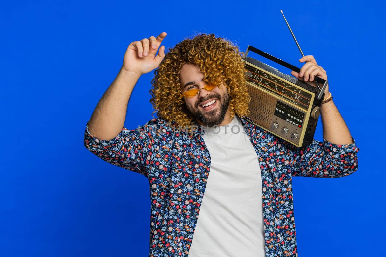 Excited man with curly hairstyle wig using retro tape record player to listen music, disco dancing favorite track, having fun, entertaining fan of vintage technologies. Guy isolated on blue background