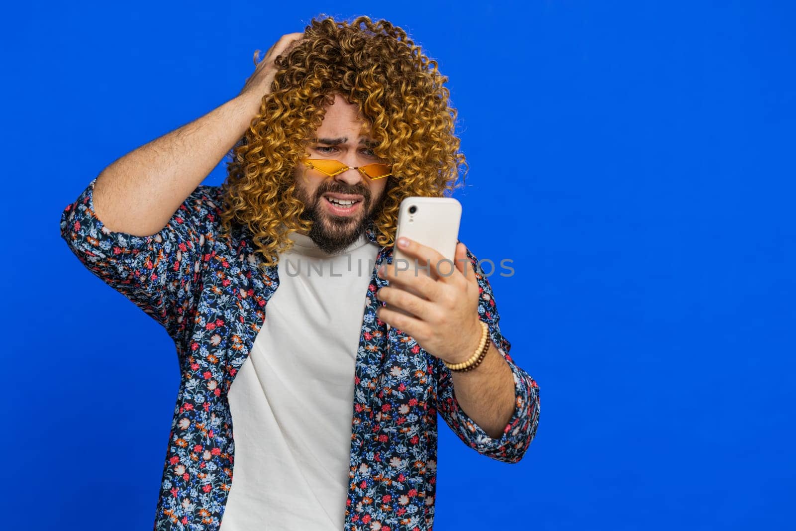 Upset sad man with curly hairstyle wig use smartphone typing browsing play game loses surprised by sudden lottery results bad fortune loss fail unlucky news. Stylish guy on blue background. Copy-space