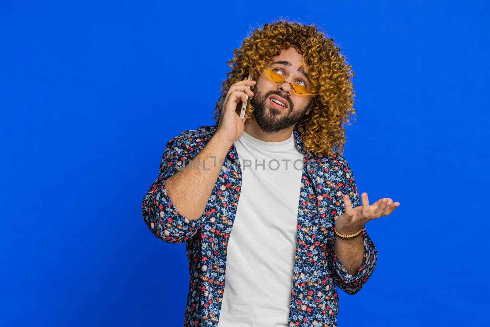 Disinterested man with curly hairstyle wig having annoyed boring talk on smartphone. Guy tired sleepy guy talking on mobile phone with friend, making online conversation on blue background. Copy-space
