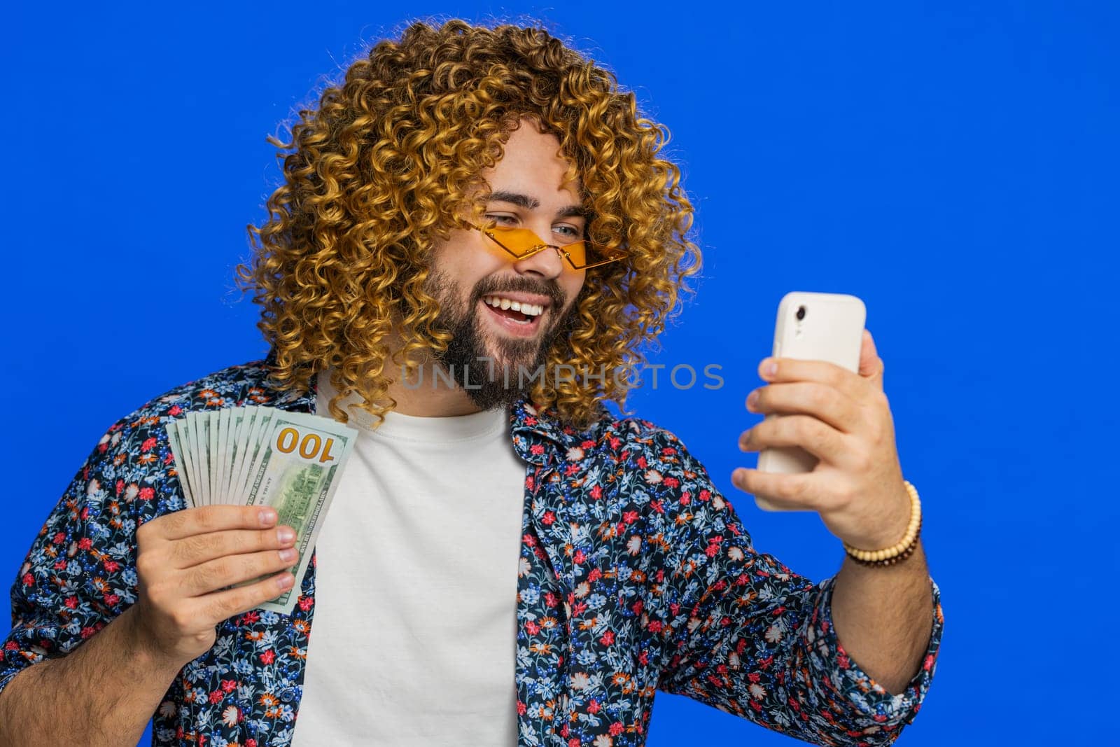 Happy rich man with curly hairstyle wig looking smartphone display sincerely rejoicing win receiving money dollar cash banknotes celebrating success victory play lottery game. Guy on blue background