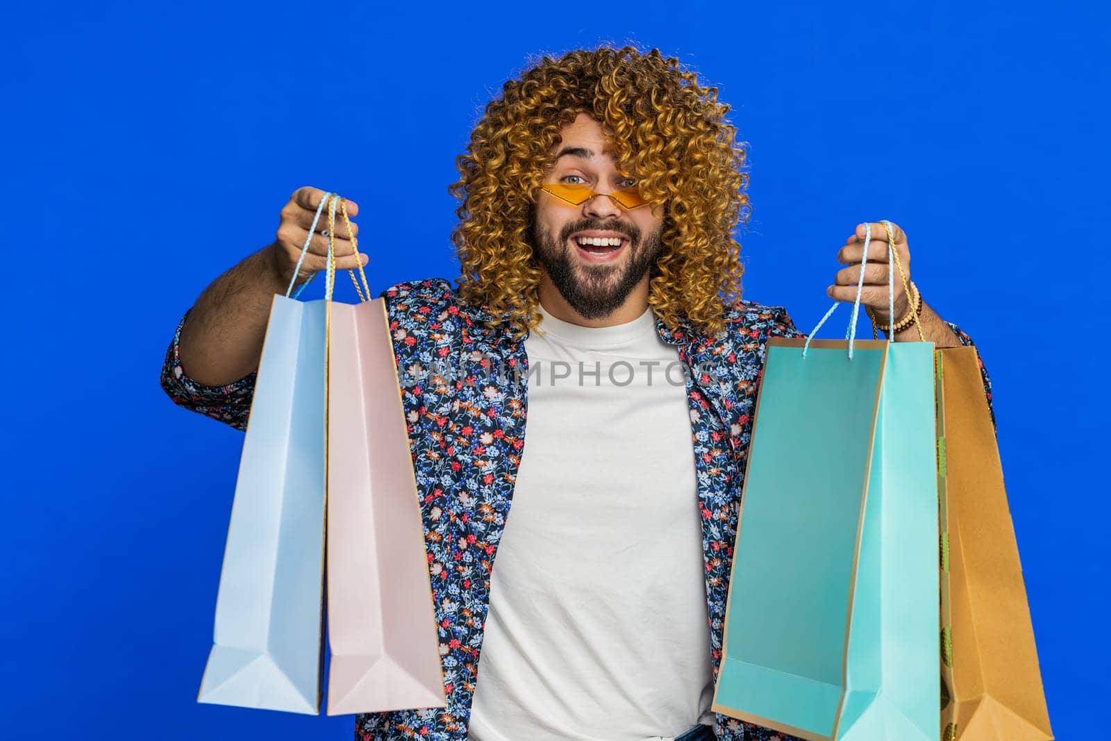 Happy Caucasian man showing shopping bags, advertising discounts, smiling looking amazed with low prices, shopping on Black Friday holidays. Stylish young guy isolated on blue background. Copy-space