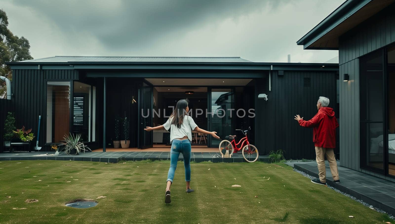 A woman and a child are playing in the backyard of a house, surrounded by green grass and plants. The facade of the building, with a roof and a lawn, overlooks the leisure event, under the cloudy sky
