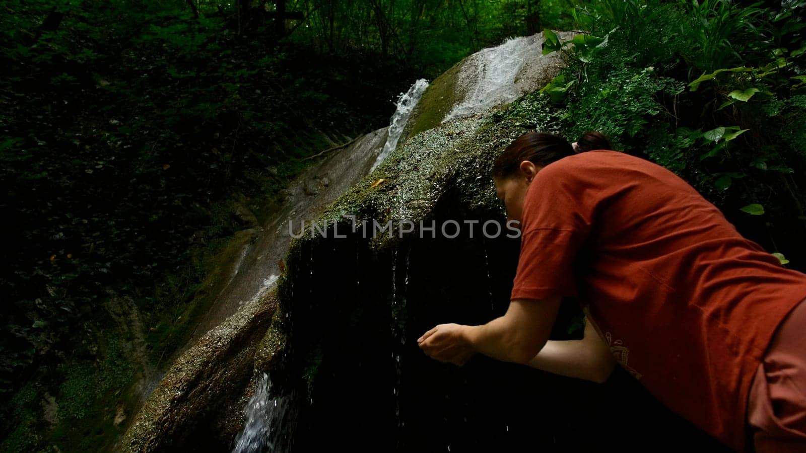 Young woman drinking water from cascade waterfall at deep tropical rain forest. Creative. Female hiker drinking water from mountainous spring. by Mediawhalestock