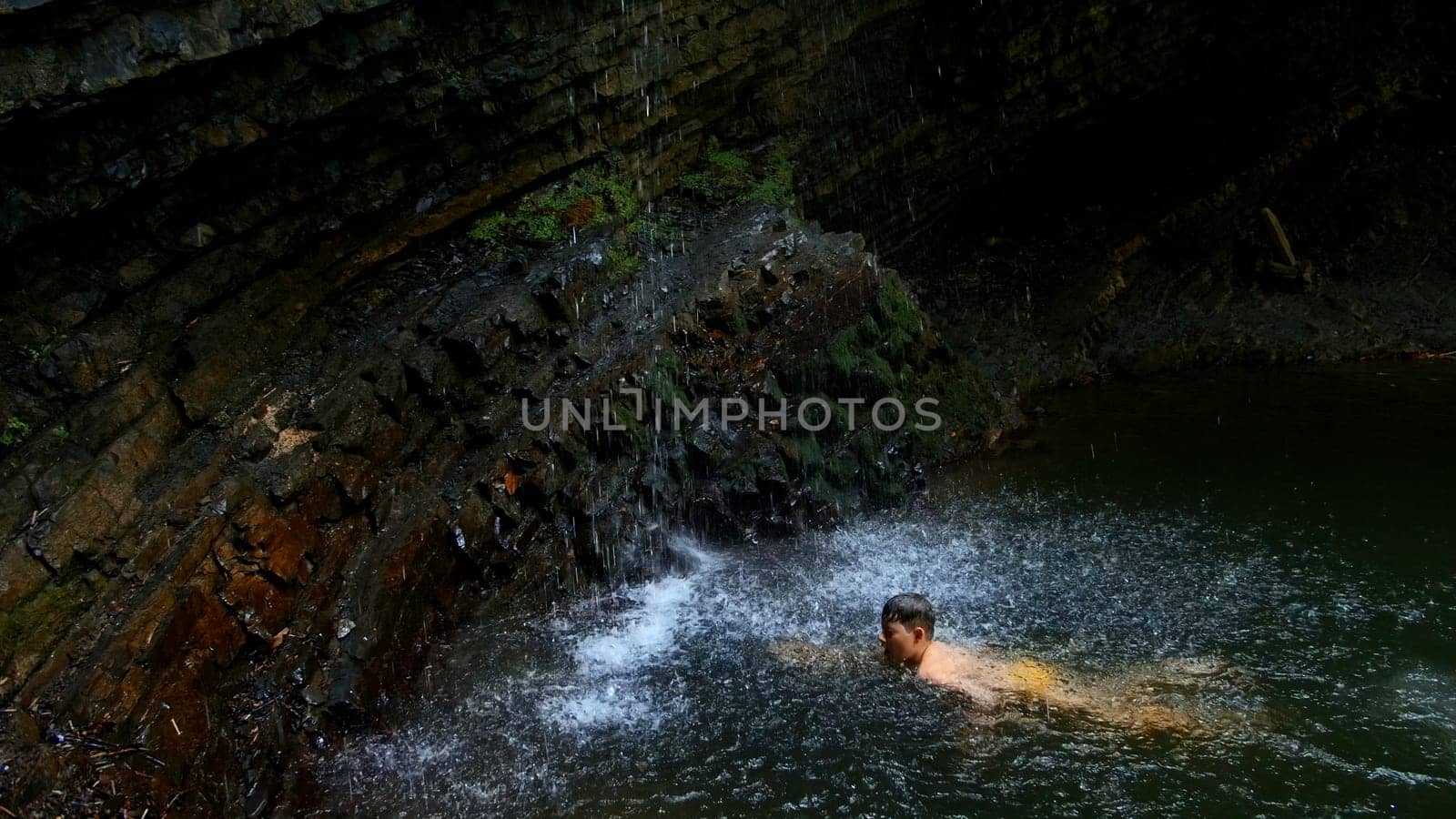 Child boy swimming in mountain pond with waterfall. Creative. Child on summertime vacation refreshing in river