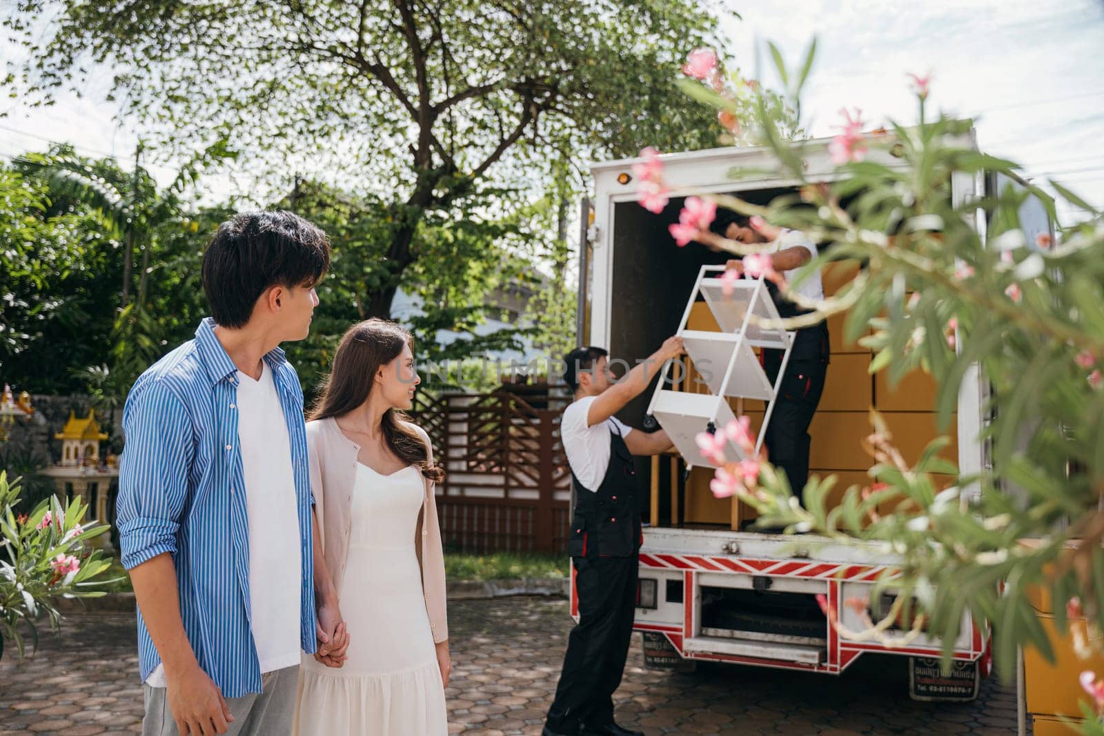 Professional movers assist a smiling newlywed couple captured in a portrait while loading furniture onto a truck. Quality service and teamwork ensure customer satisfaction. Moving Day Concept by Sorapop