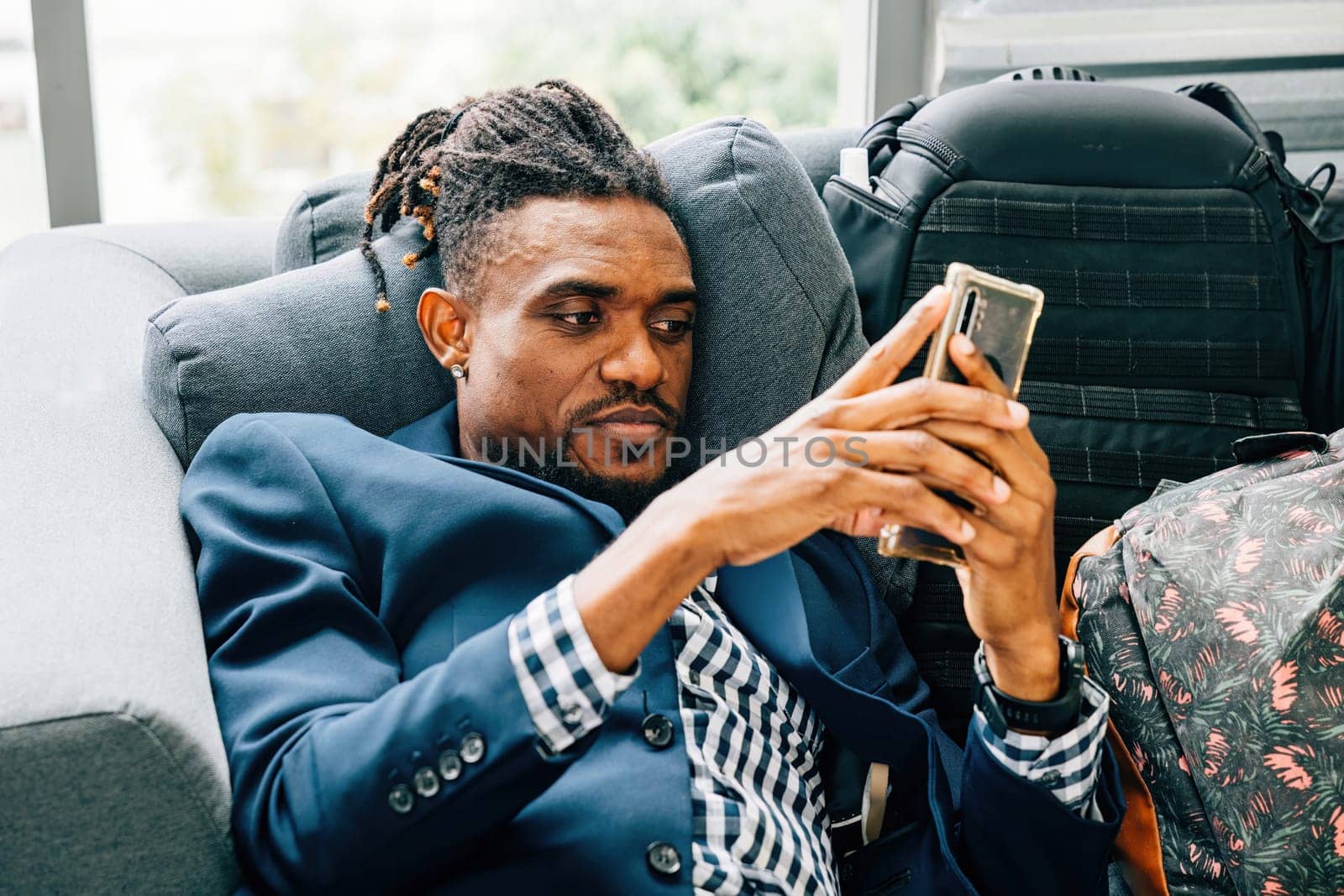 A cheerful black businessman is seen at home, using his smartphone to check social networks, read messages, and browse the internet while sitting on a cozy sofa in a living room. relax time