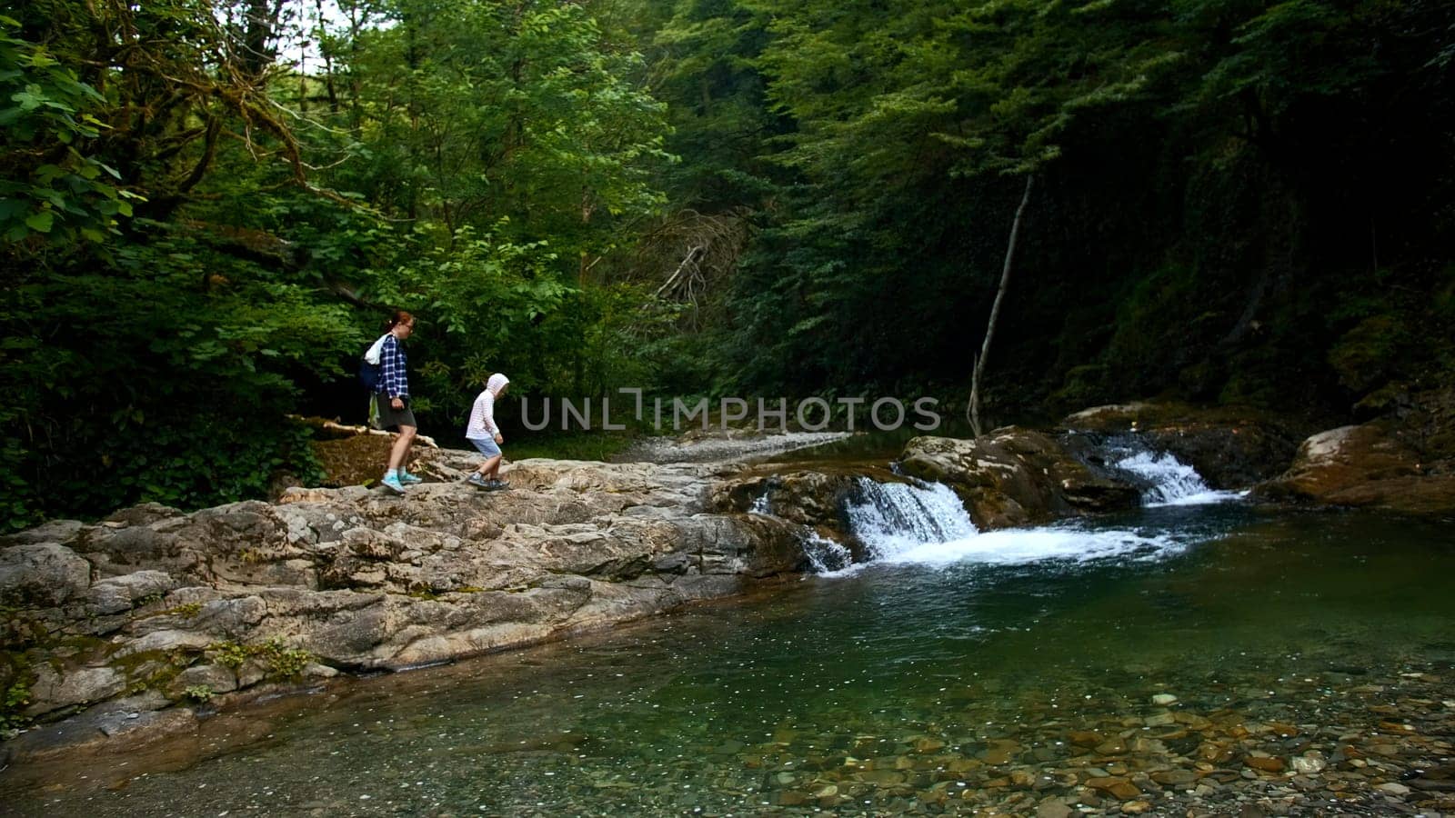 Woman and boy walking along natural pond with small waterfall, summer time. Creative. Travelling in jungles