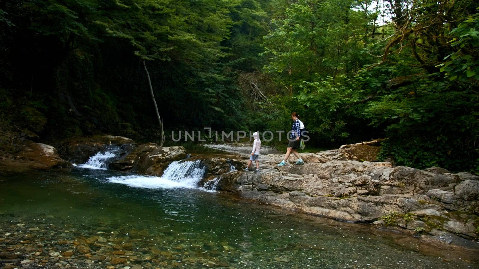 Woman and boy walking along natural pond with small waterfall, summer time. Creative. Travelling in jungles. by Mediawhalestock