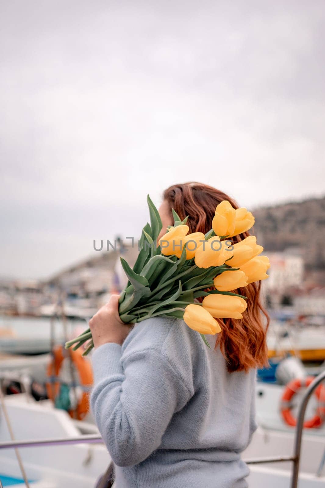 Woman holds yellow tulips in harbor with boats docked in the background., overcast day, yellow sweater, mountains by Matiunina