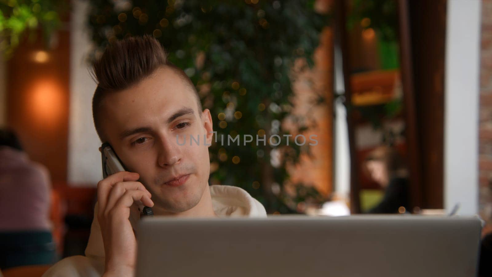 Young man is working on laptop in cafe and talking on phone. Stock footage. Young freelancer is working on laptop and answering calls. Remote work with laptop and cafe calls.