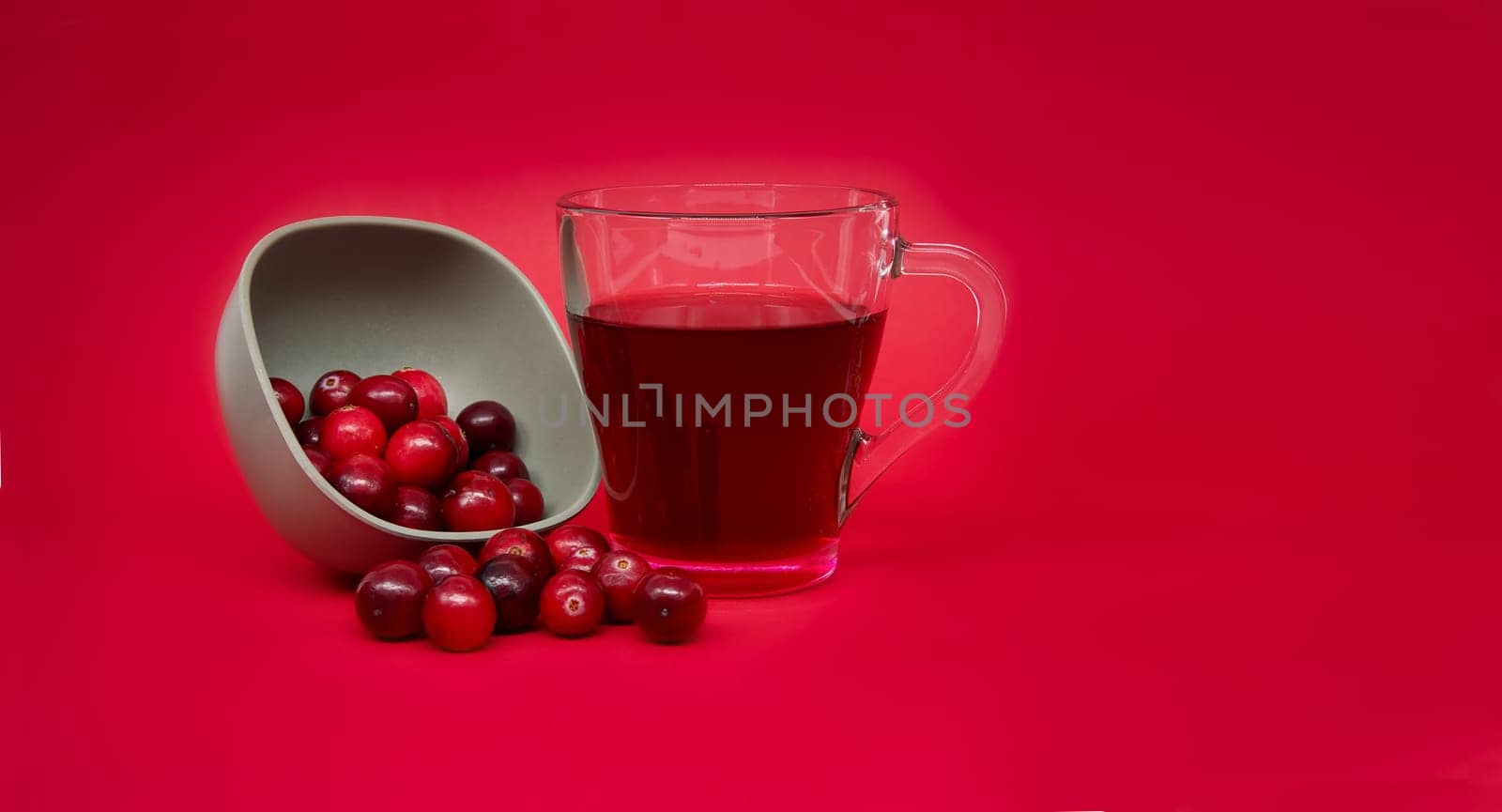 Grey ceramic bowl full of red cranberries and transparent cup containing a dark red cranberry juice against a vibrant red background