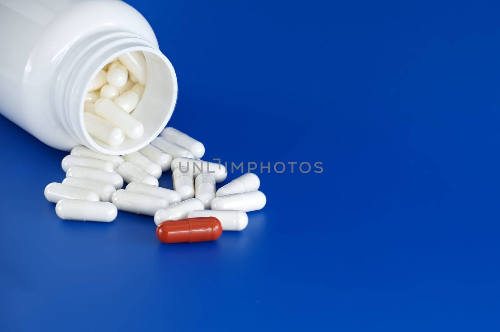 Overturned white medicine bottle with its contents white pills or capsules, spilled out onto a blue background. Among these white pills, a solitary red pill stands out