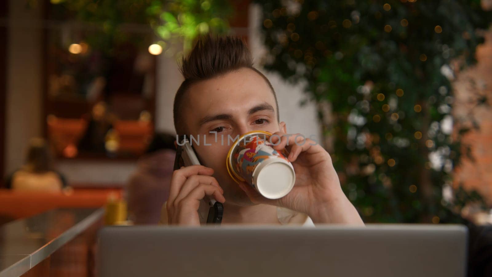 Young man is working on laptop in cafe and talking on phone. Stock footage. Young freelancer is working on laptop and answering calls. Remote work with laptop and cafe calls.
