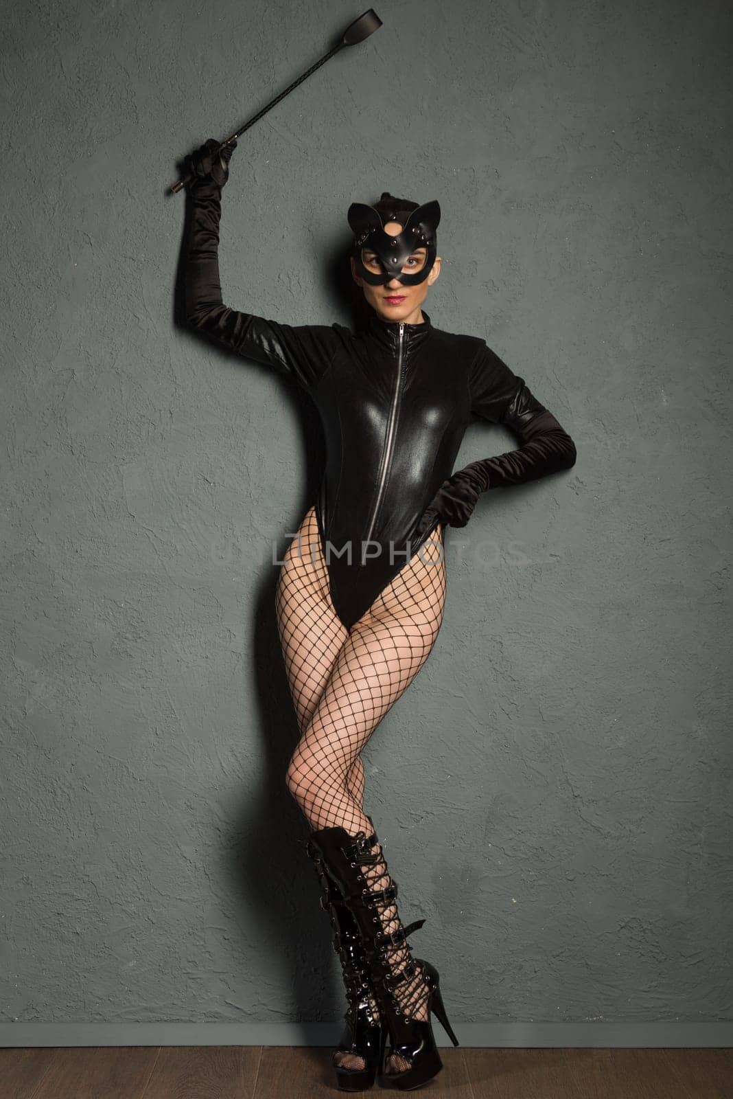 Adult sex games. Beautiful dominant brunette vamp mistress girl in latex body, gloves and bdsm black leather fetish cat mask posing with riding crop. - image