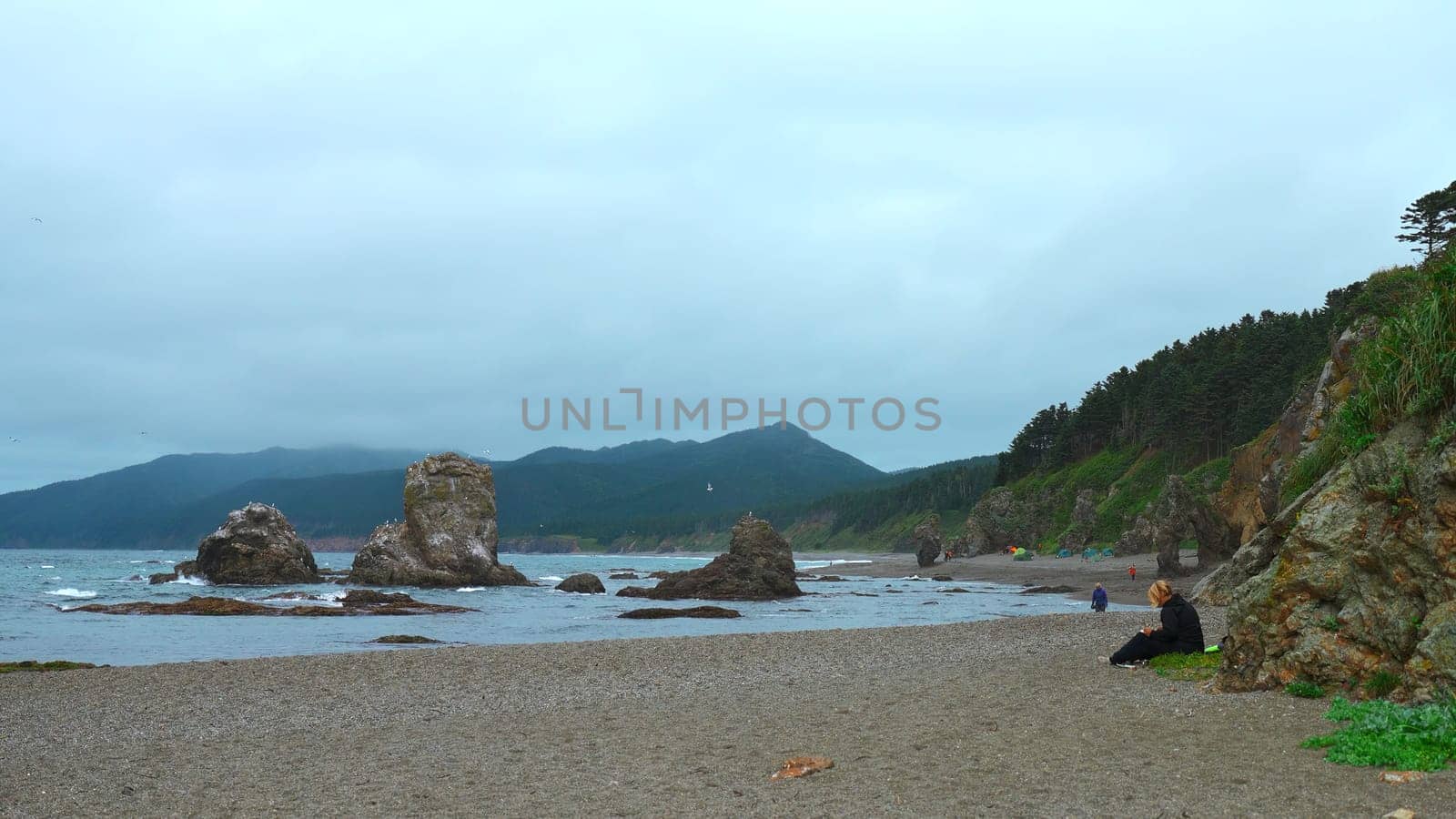 People relax on rocky coast with mountains on cloudy day. Clip. Beautiful coast with rocks and rocks sticking out on shore. People relax on beautiful coast with rocks and mountains.
