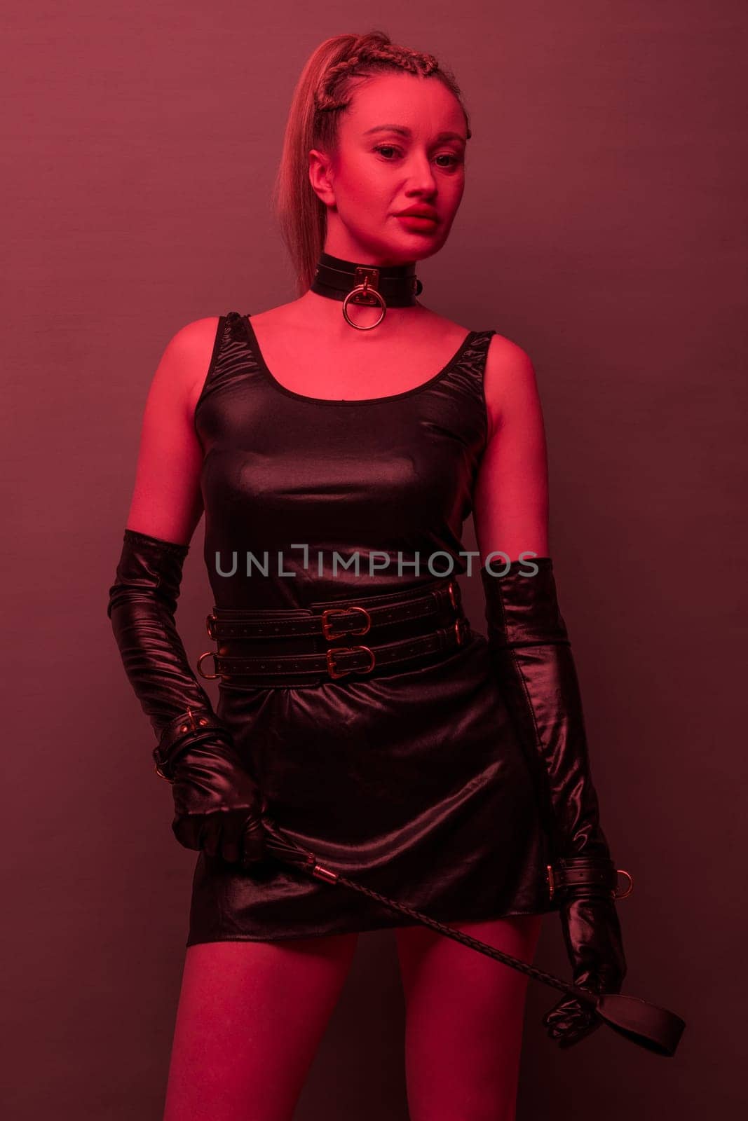 Beautiful dominant brunette mistress woman in latex dress and gloves posing with riding crop on red light backgound by zartarn