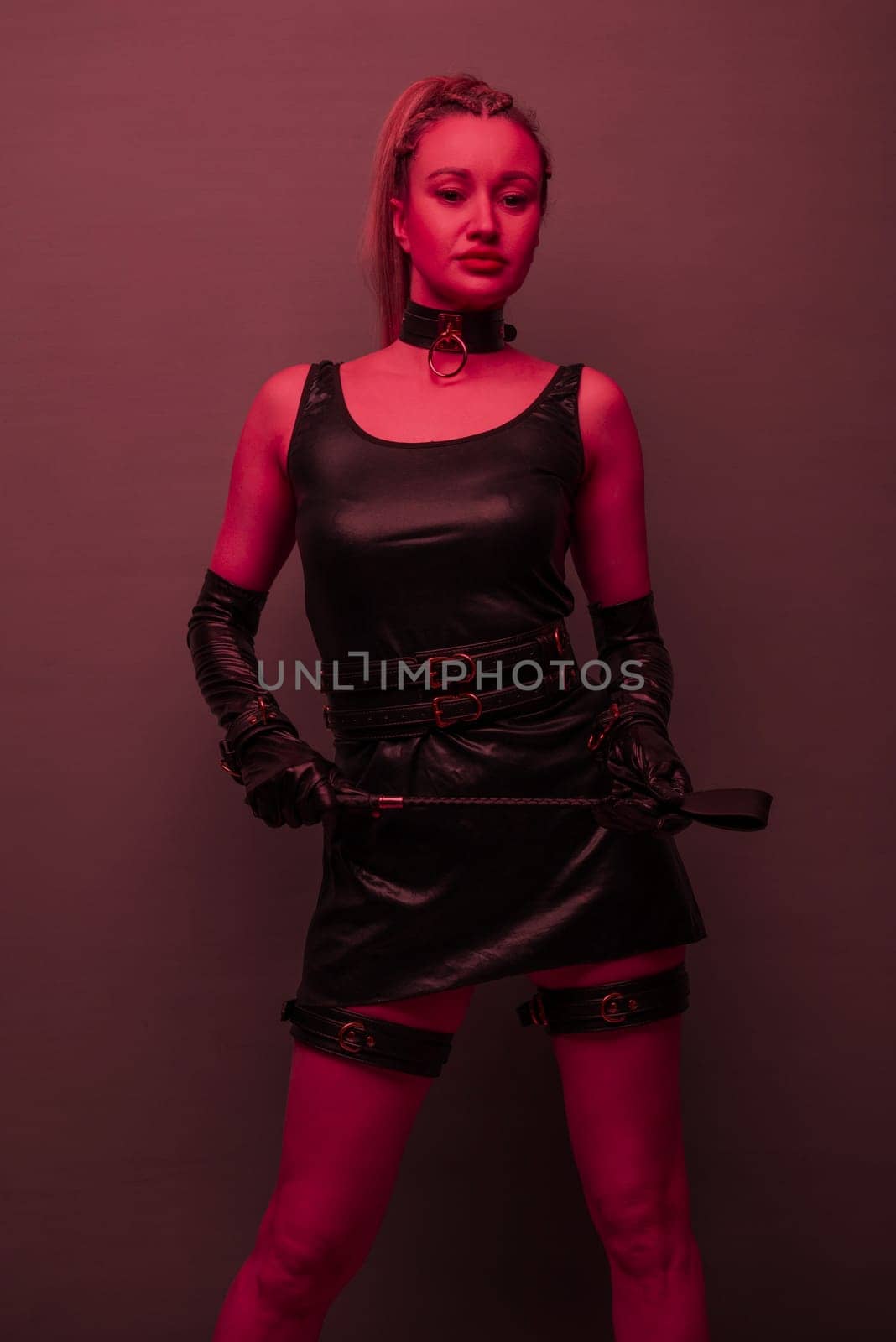 Beautiful dominant brunette mistress woman in latex dress and gloves posing with riding crop on red light backgound